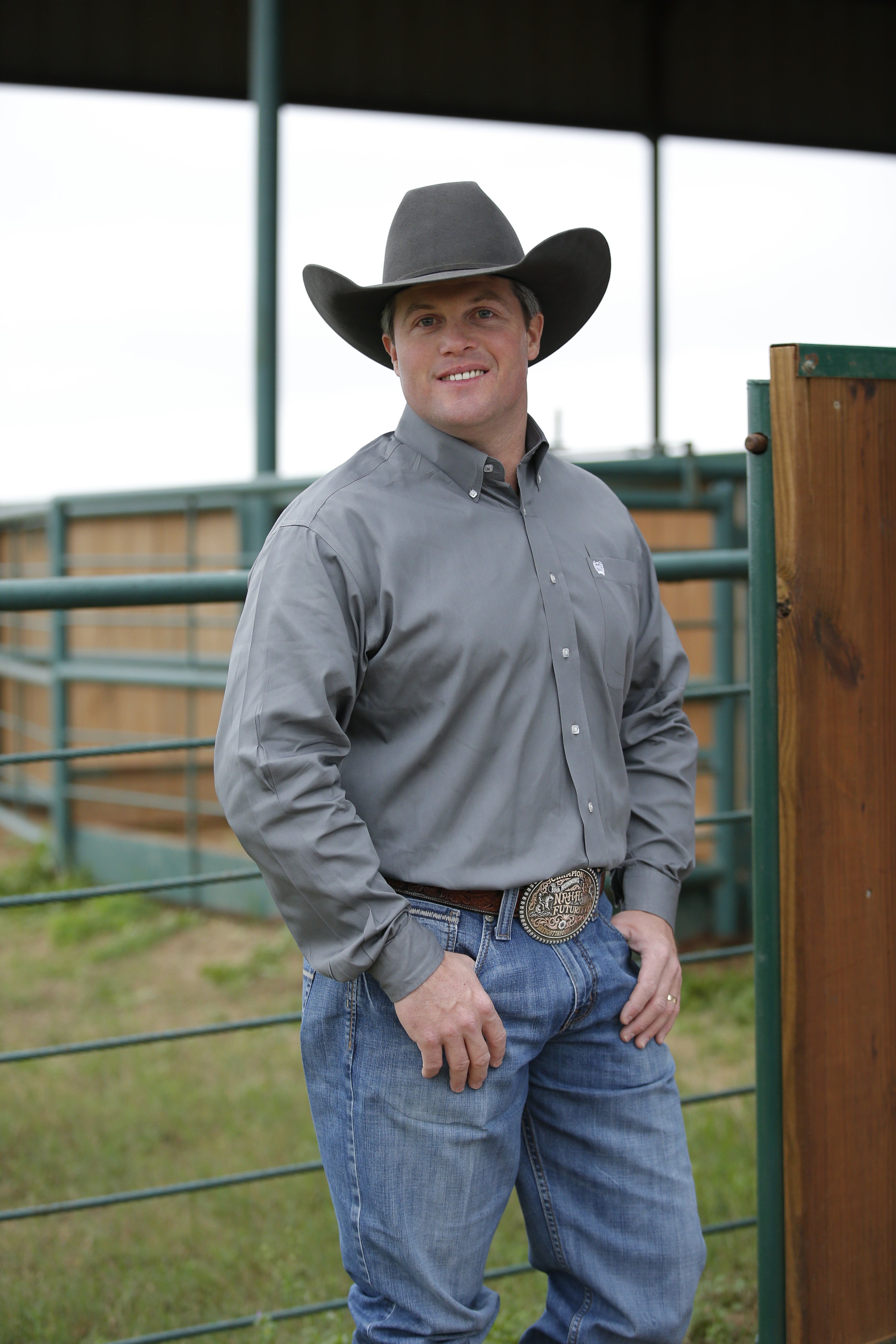 A man wearing a long-sleeve grey button-down shirt and blue jeans, with a belt and buckle visible standing outside