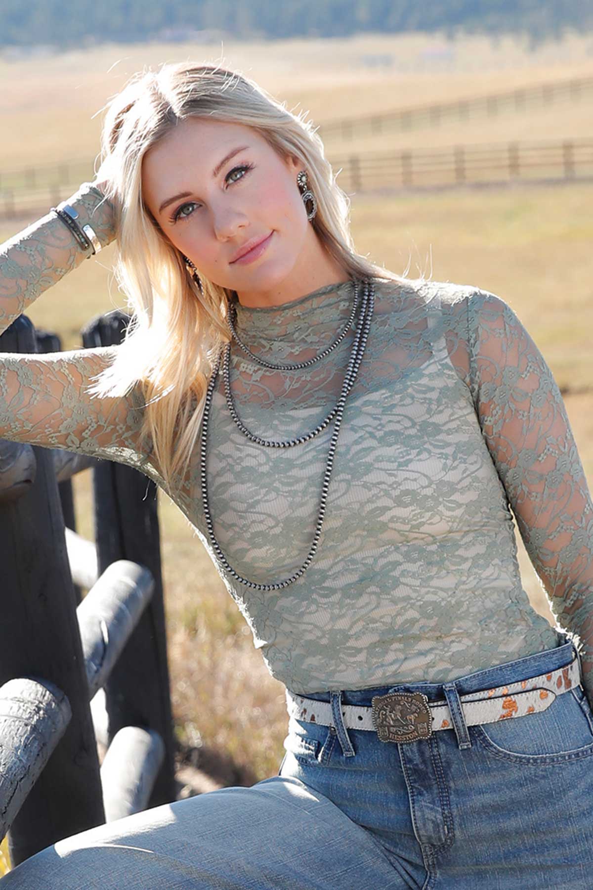Woman wearing a sheer sage green lace long-sleeve top with layered silver necklaces, light-wash jeans, and a decorative belt buckle, leaning against a wooden fence outdoors