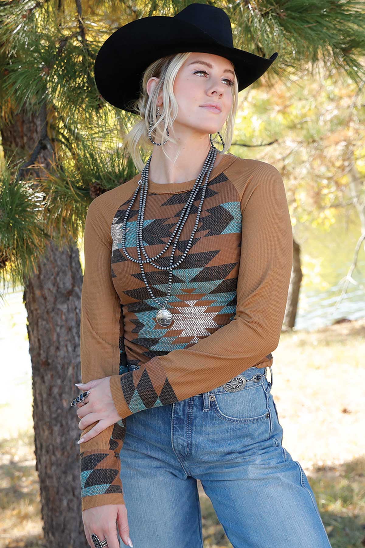 Woman wearing a brown sweater with an aztec pattern, blue jeans, and a black cowboy hat in a natural setting.