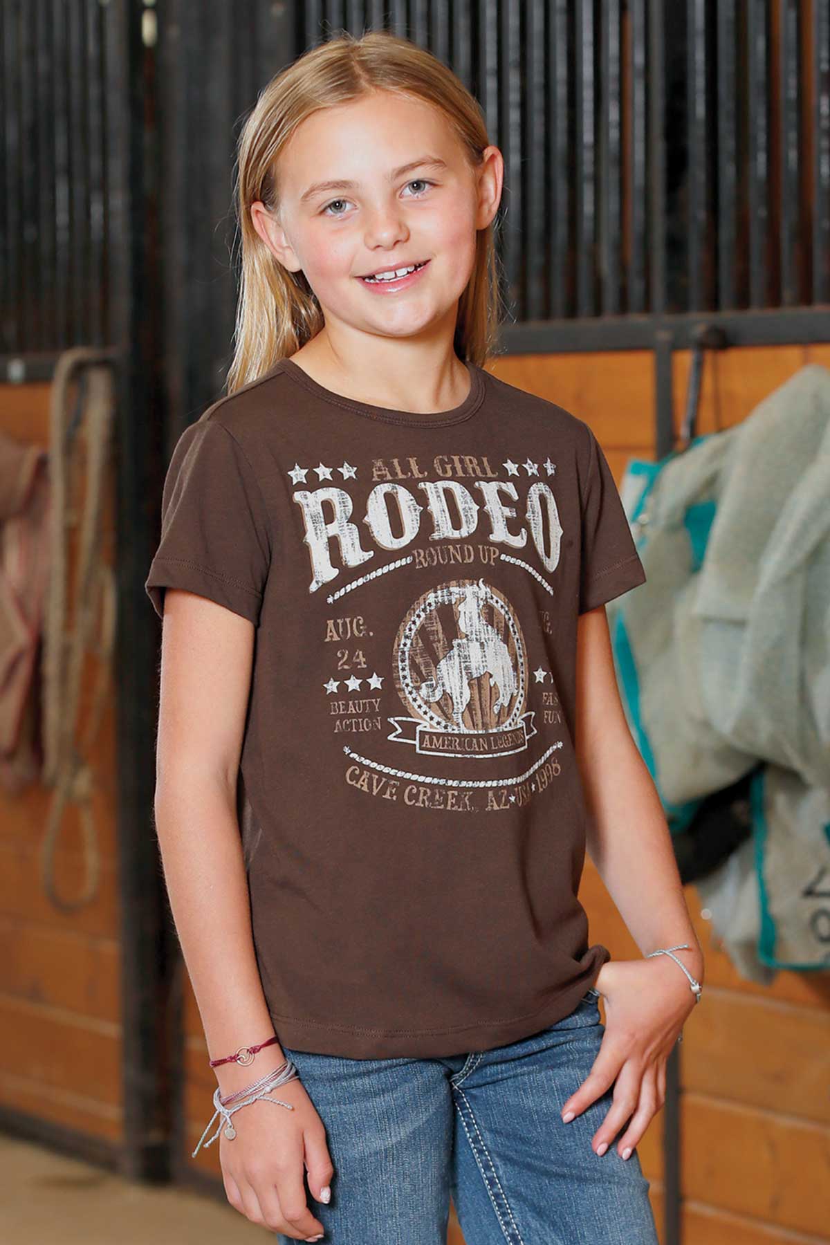 Young girl wearing a brown 'All Girl Rodeo' t-shirt in an indoor setting.