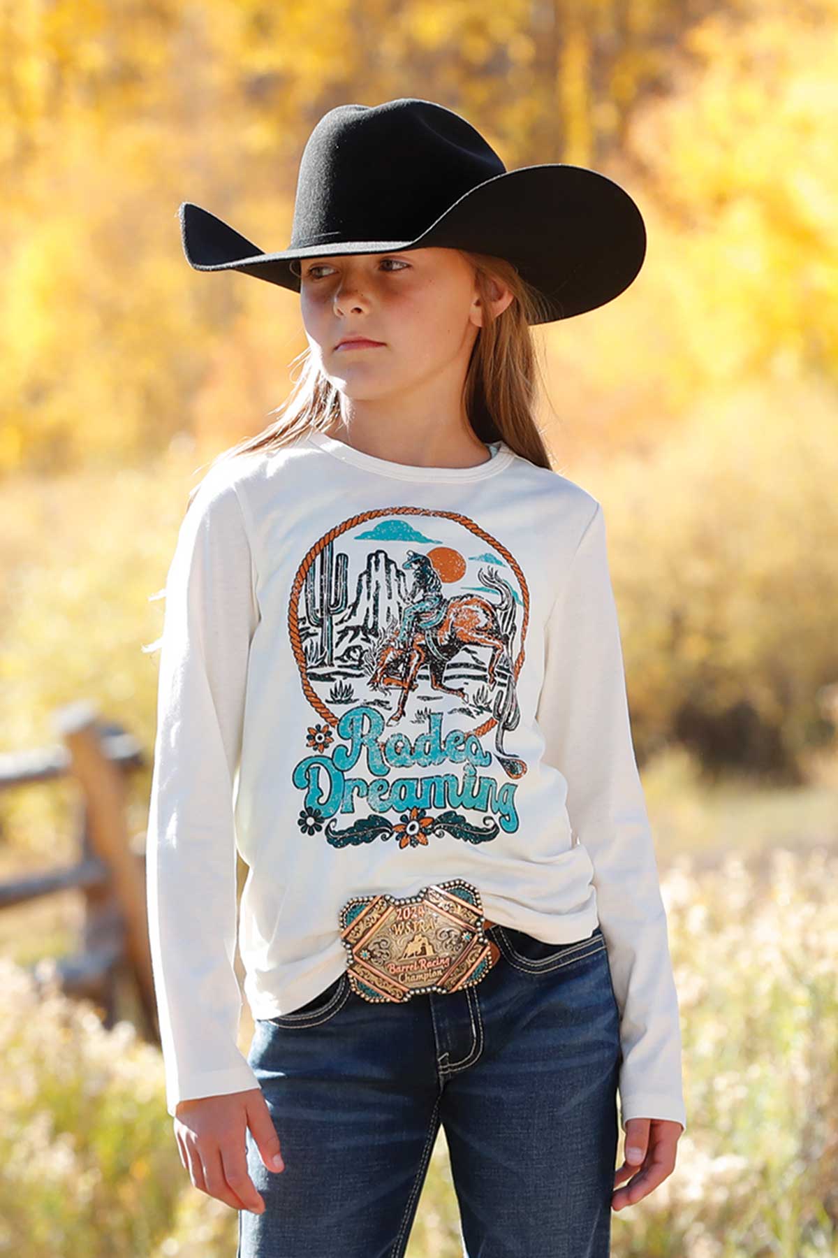 Young girl outdoors wearing a white long sleeve shirt with a colorful western graphic and the text 'Rodeo Dreamin’ on the front. She has on dark jeans, a belt with an ornate buckle, and a black cowboy hat. Autumn foliage is visible in the background