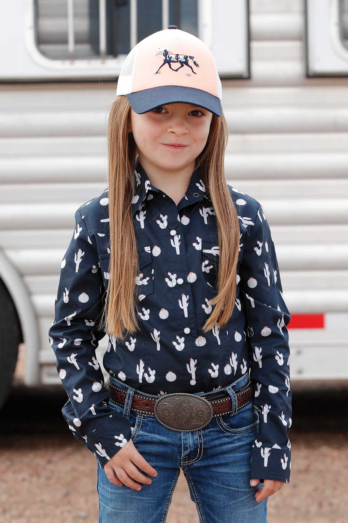 Child wearing a navy patterned shirt and pink cap in front of a trailer.