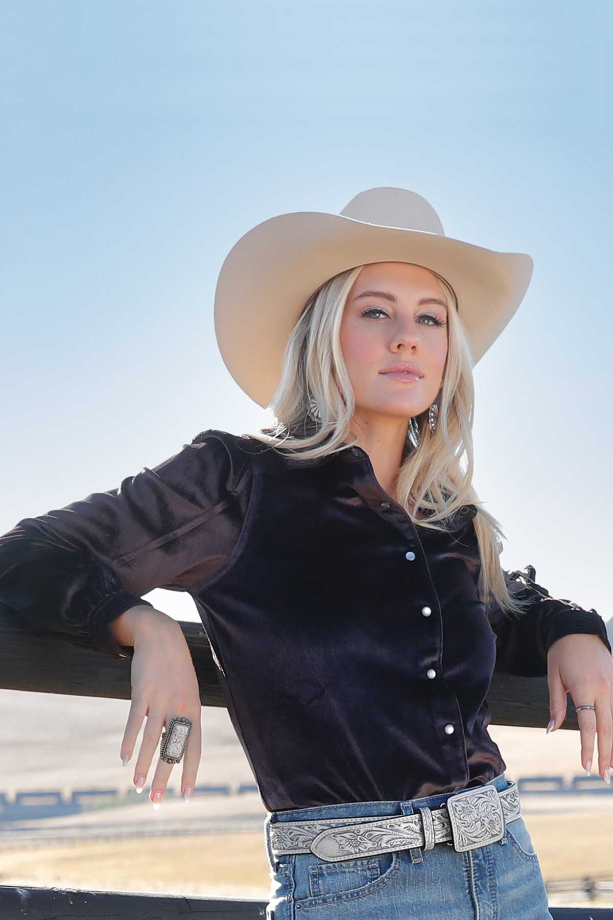 Woman wearing a black long-sleeve western shirt, light-colored cowboy hat, silver belt buckle, and blue jeans, leaning against a wooden fence outdoors