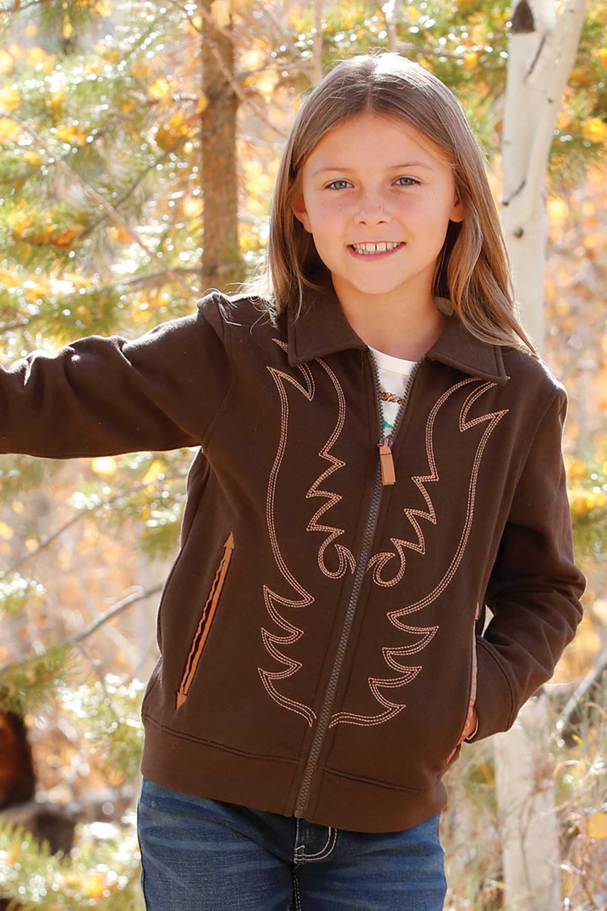 Girl wearing a brown zip-up jacket with tan western-style embroidery on the front, paired with jeans, standing outdoors among trees with autumn leaves and smiling