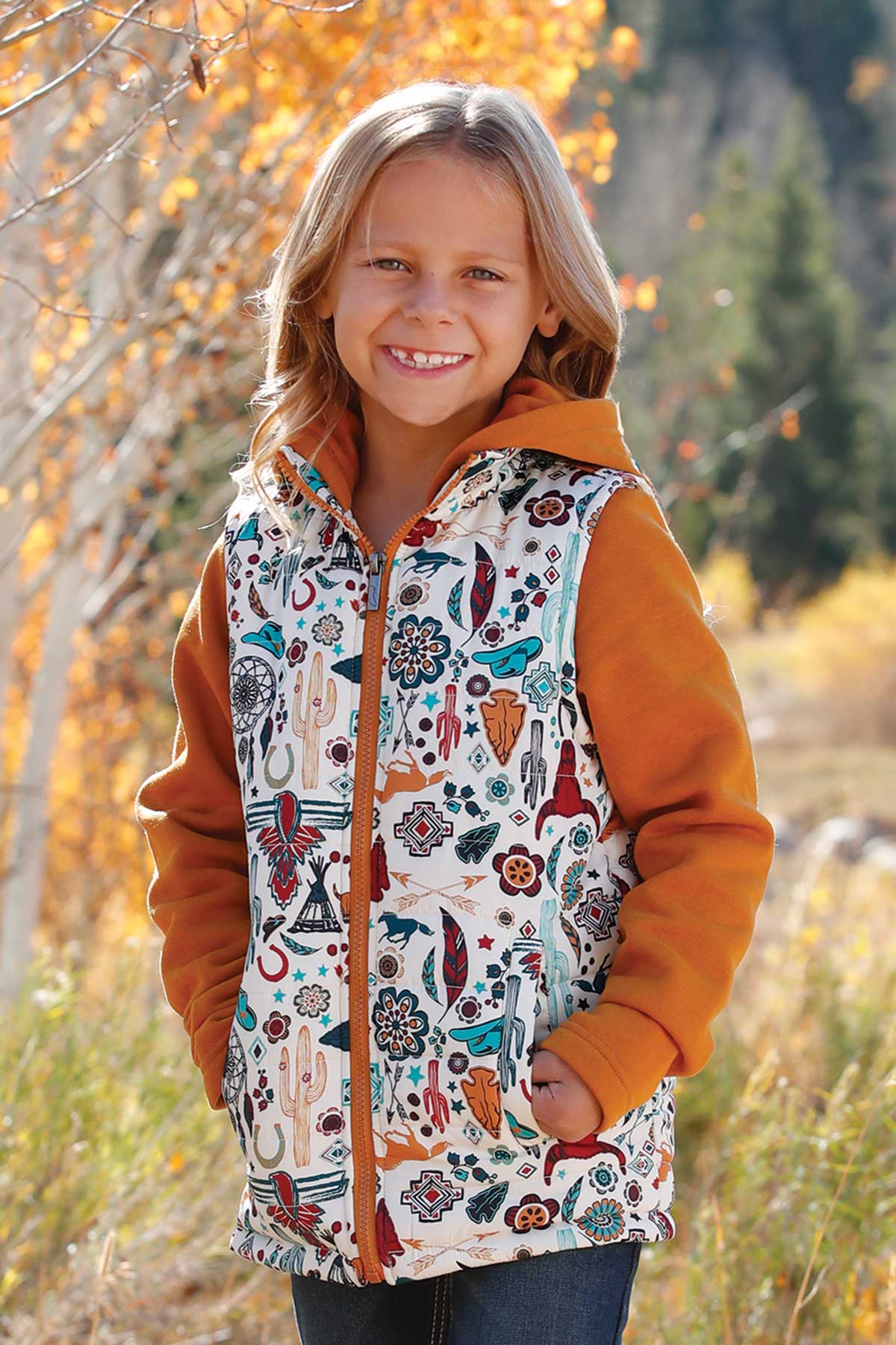Girl wearing a rust orange hoodie layered with a cream zip-up vest featuring colorful western-inspired prints, standing outdoors with autumn trees in the background and smiling