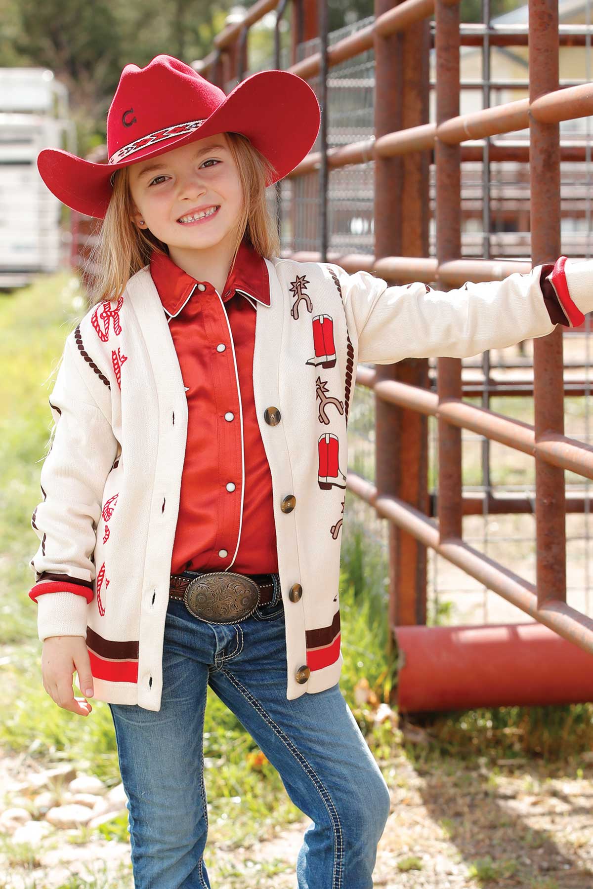Girl wearing a red cowboy hat, red button-up shirt, and cream cardigan with western embroidery, paired with blue jeans and a decorative belt buckle, smiling and standing by a metal fence