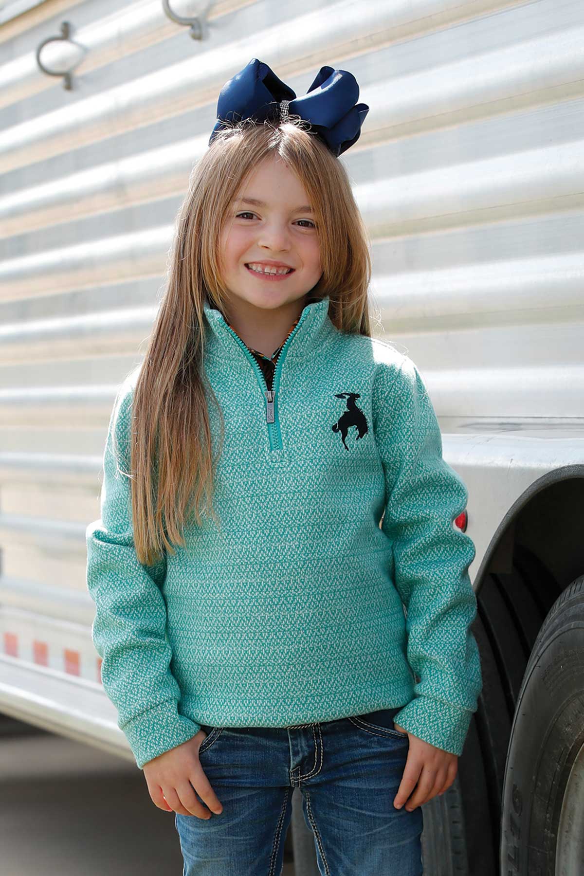 Girl wearing a turquoise quarter-zip pullover with a black rodeo rider logo on the chest, paired with jeans and a large navy bow in her hair, smiling while standing next to a trailer