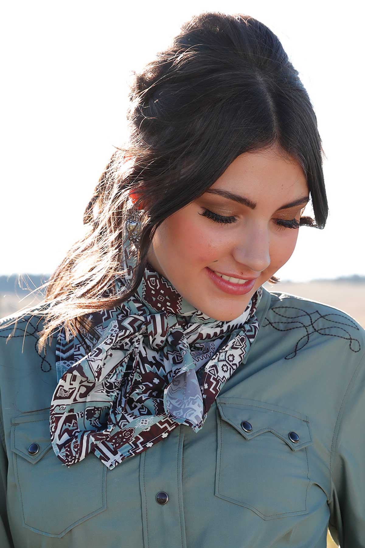 Woman wearing a light green western shirt with black embroidery and a patterned scarf tied around the neck, smiling and looking down