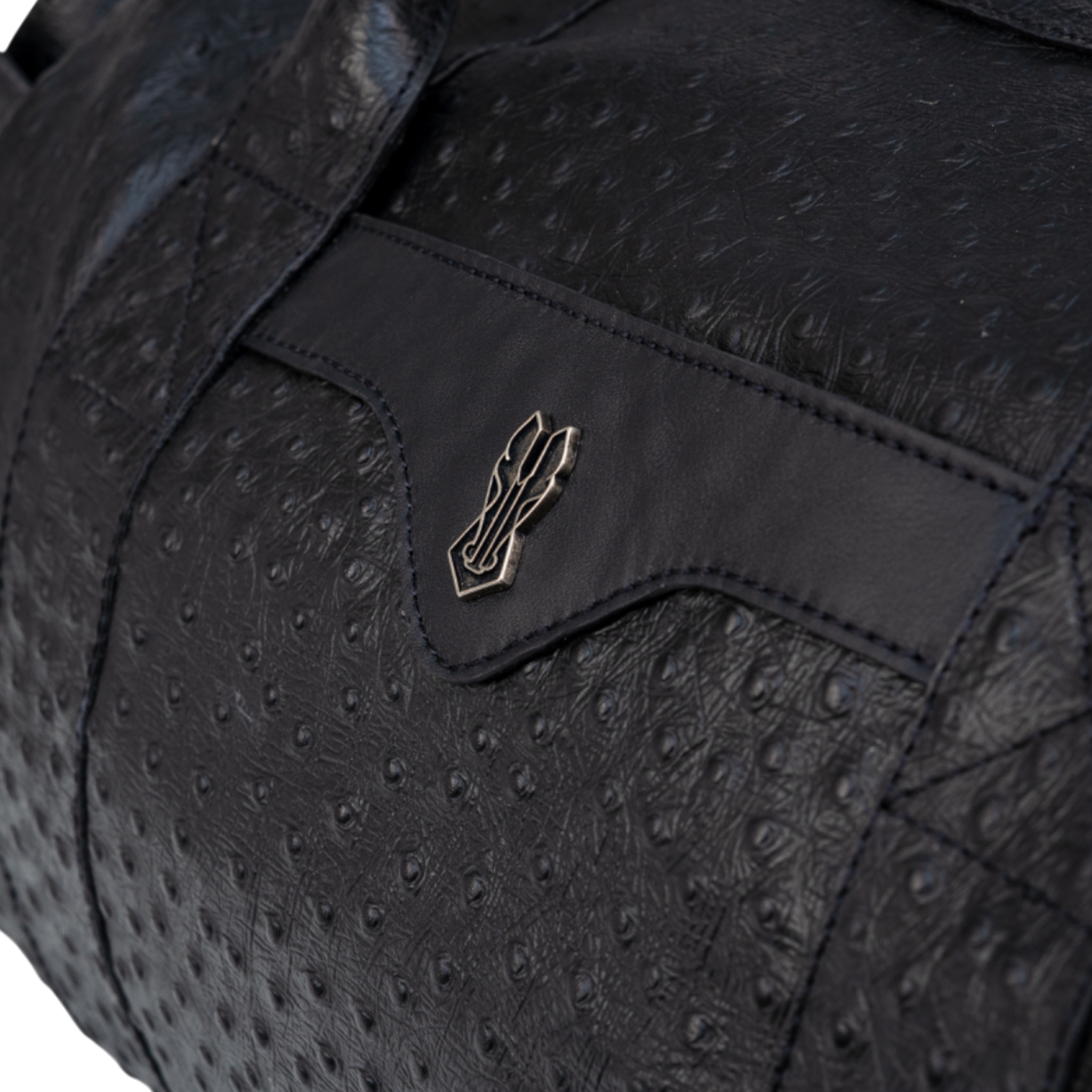 Close-up of a black bag featuring a textured ostrich-print leather pattern and a smooth leather accent with a small metallic emblem in the center