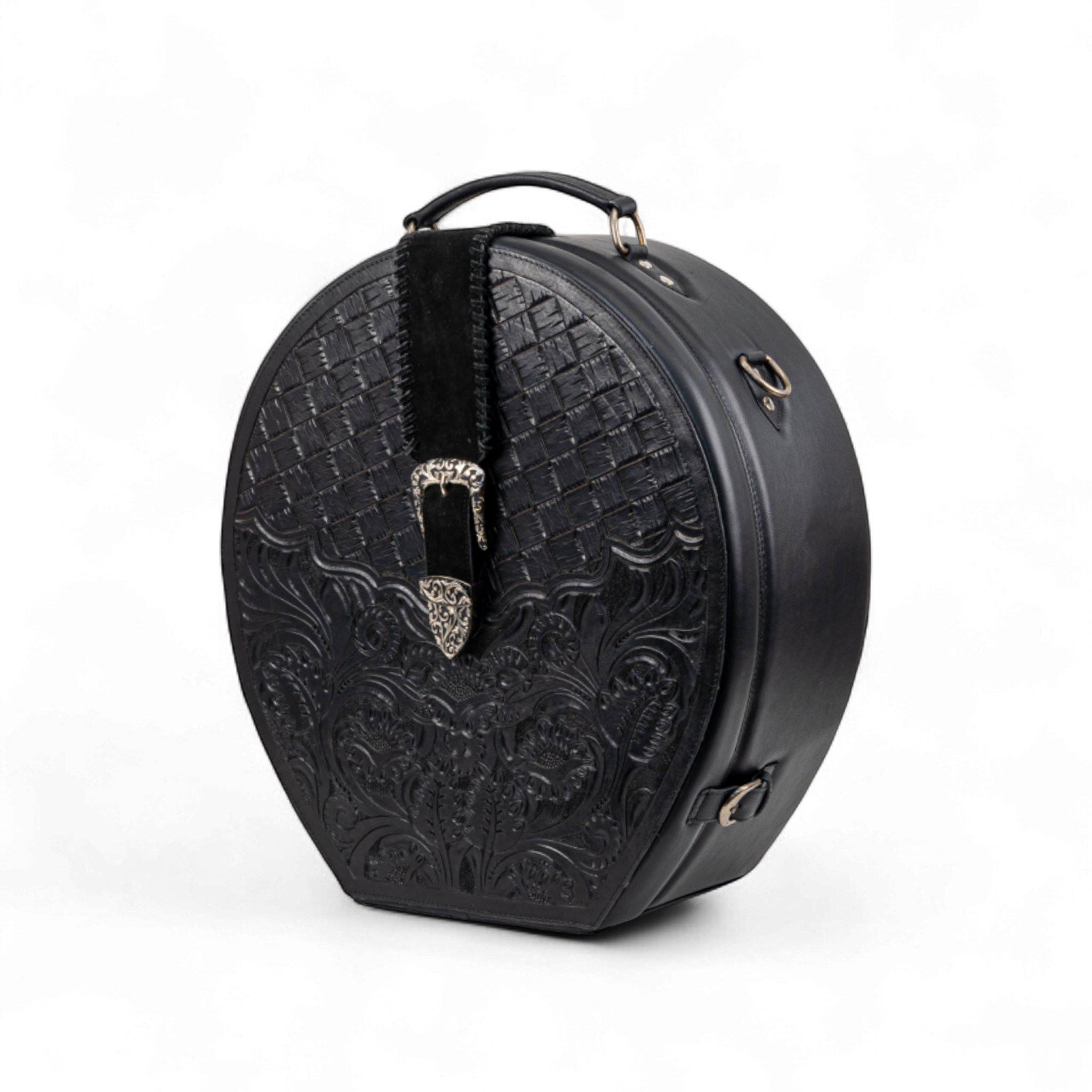 Black tooled leather hat case with a basket-weave and floral design on the front, accented by a silver buckle on a suede strap. The case has a structured round shape, side hardware rings, and a detachable shoulder strap