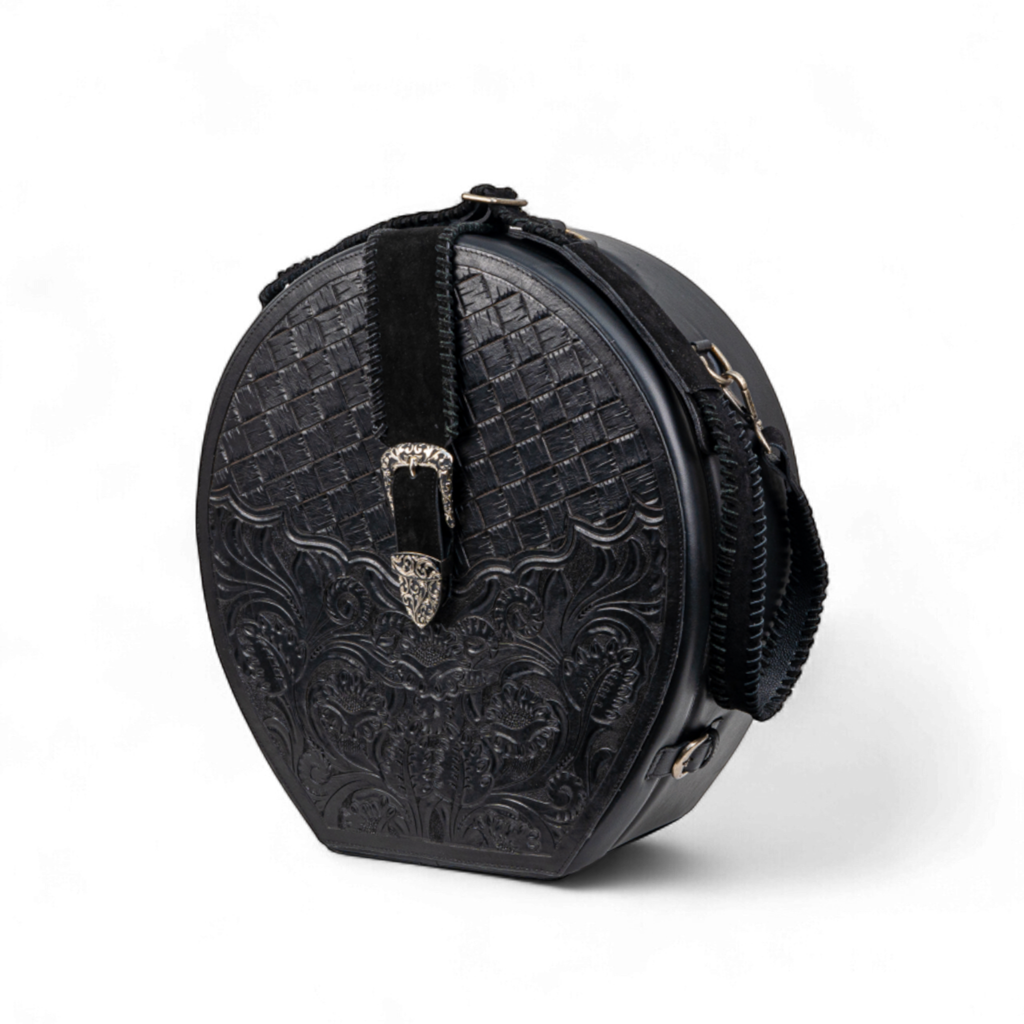 Black tooled leather hat case with a basket-weave and floral design on the front, accented by a silver buckle on a suede strap. The case has a structured round shape, side hardware rings, and a detachable shoulder strap