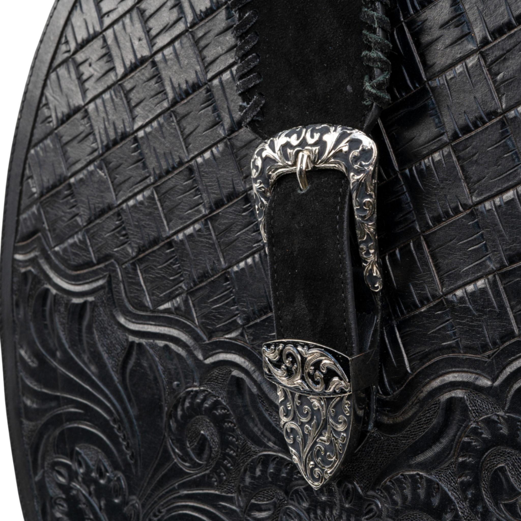 Close-up of an ornate silver buckle on a black textured leather surface, featuring a mix of basket-weave and floral tooling patterns