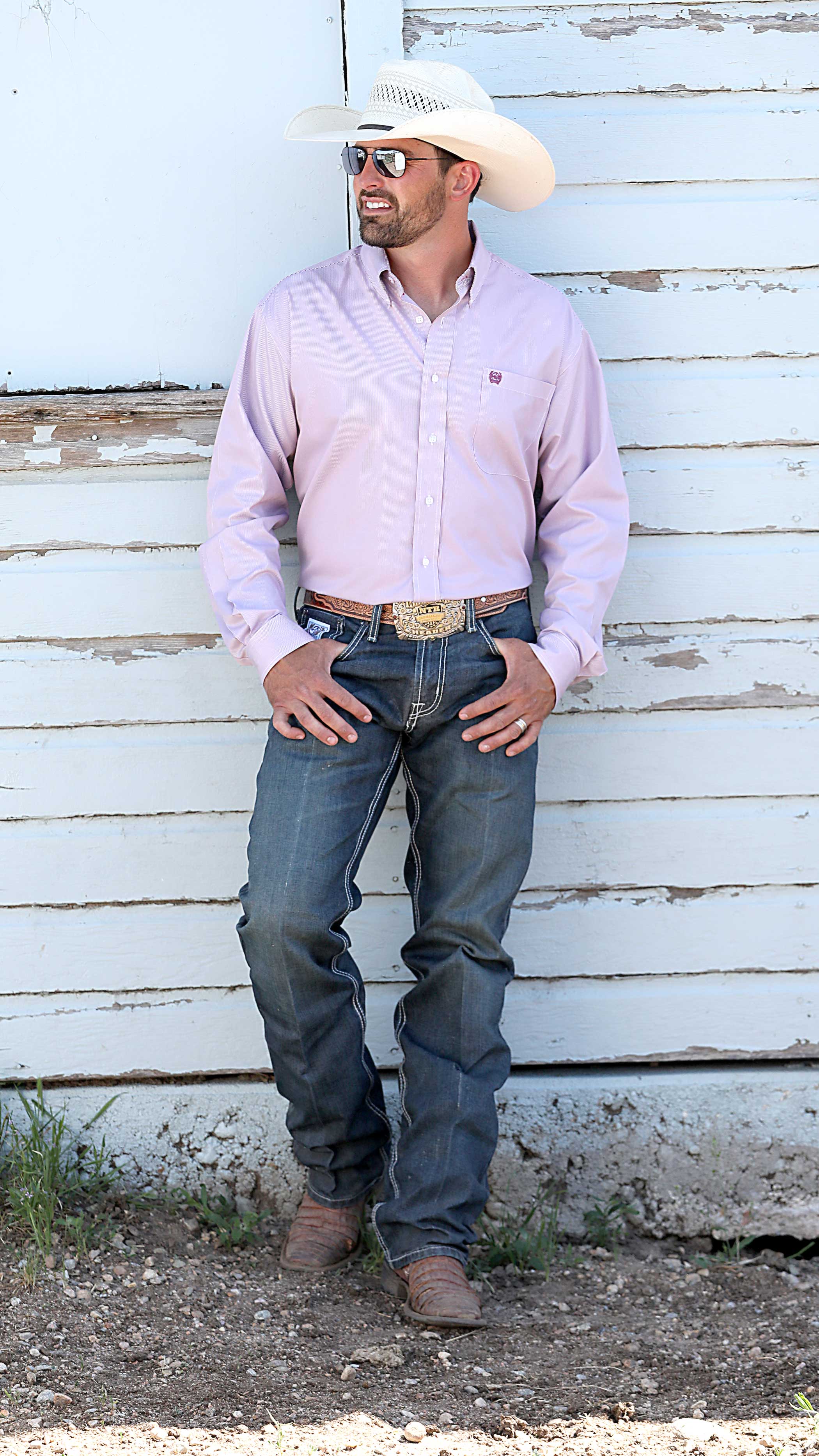 Man wearing a white cowboy hat, light pink button-down shirt, blue jeans with a belt buckle, and cowboy boots, standing casually against a white wooden wall with hands hooked in his belt loops