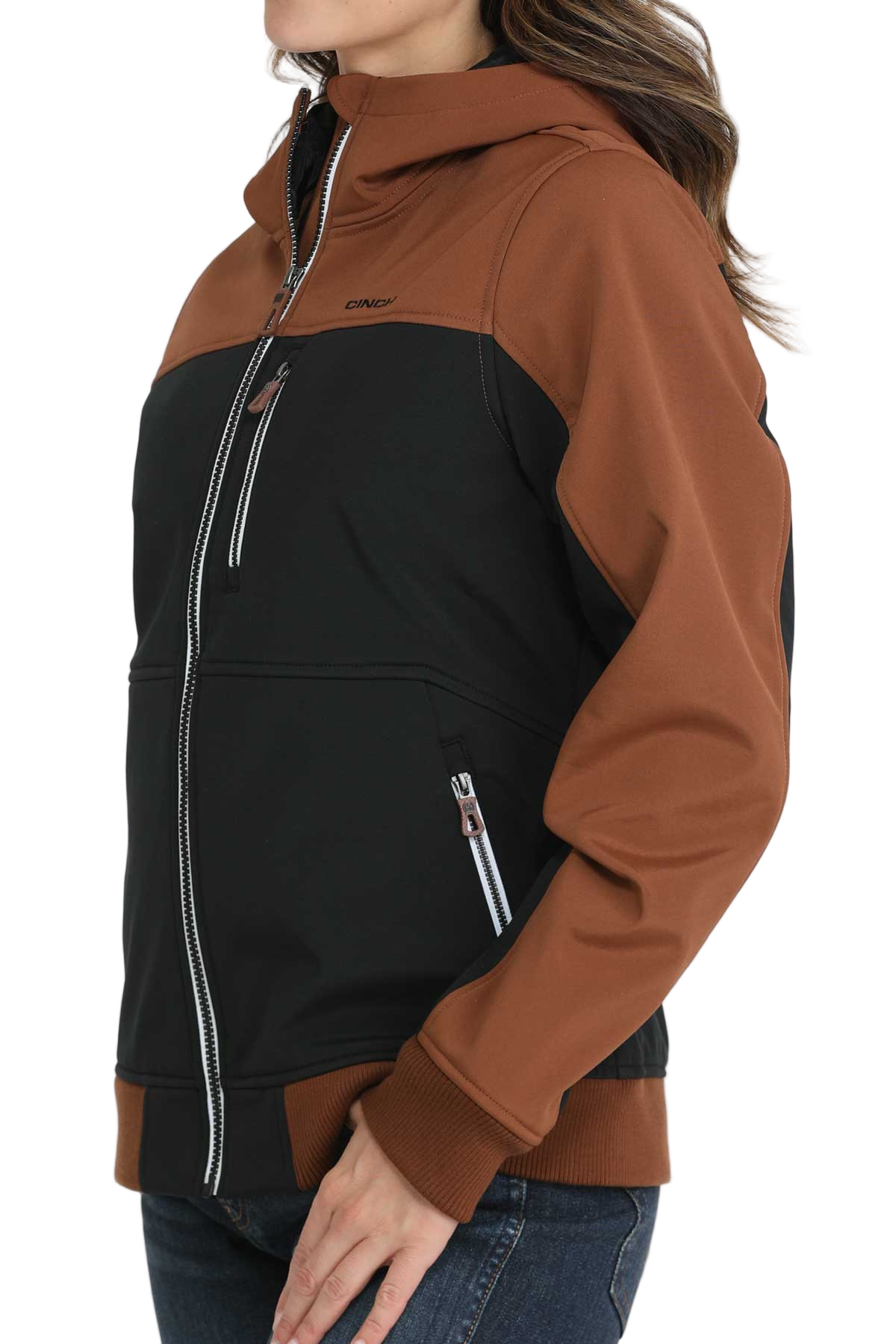 Woman wearing a two-tone hooded jacket with a brown upper section and sleeves and a black lower body. The jacket features a full front zipper, white contrast stitching, and zippered pockets. She pairs it with blue jeans.