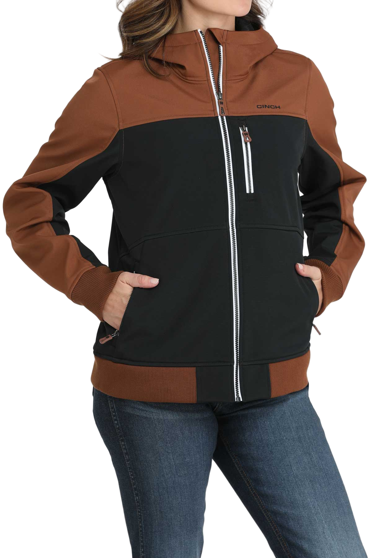Woman wearing a two-tone hooded jacket with a brown upper section and sleeves and a black lower body. The jacket features a full front zipper, white contrast stitching, and zippered pockets. She pairs it with blue jeans.