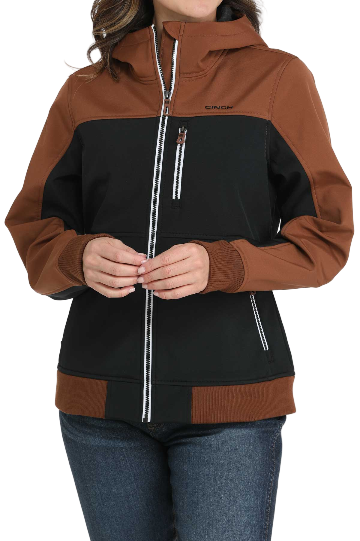 Woman wearing a two-tone hooded jacket with a brown upper section and sleeves and a black lower body. The jacket features a full front zipper, white contrast stitching, and zippered pockets. She pairs it with blue jeans, standing with hands clasped