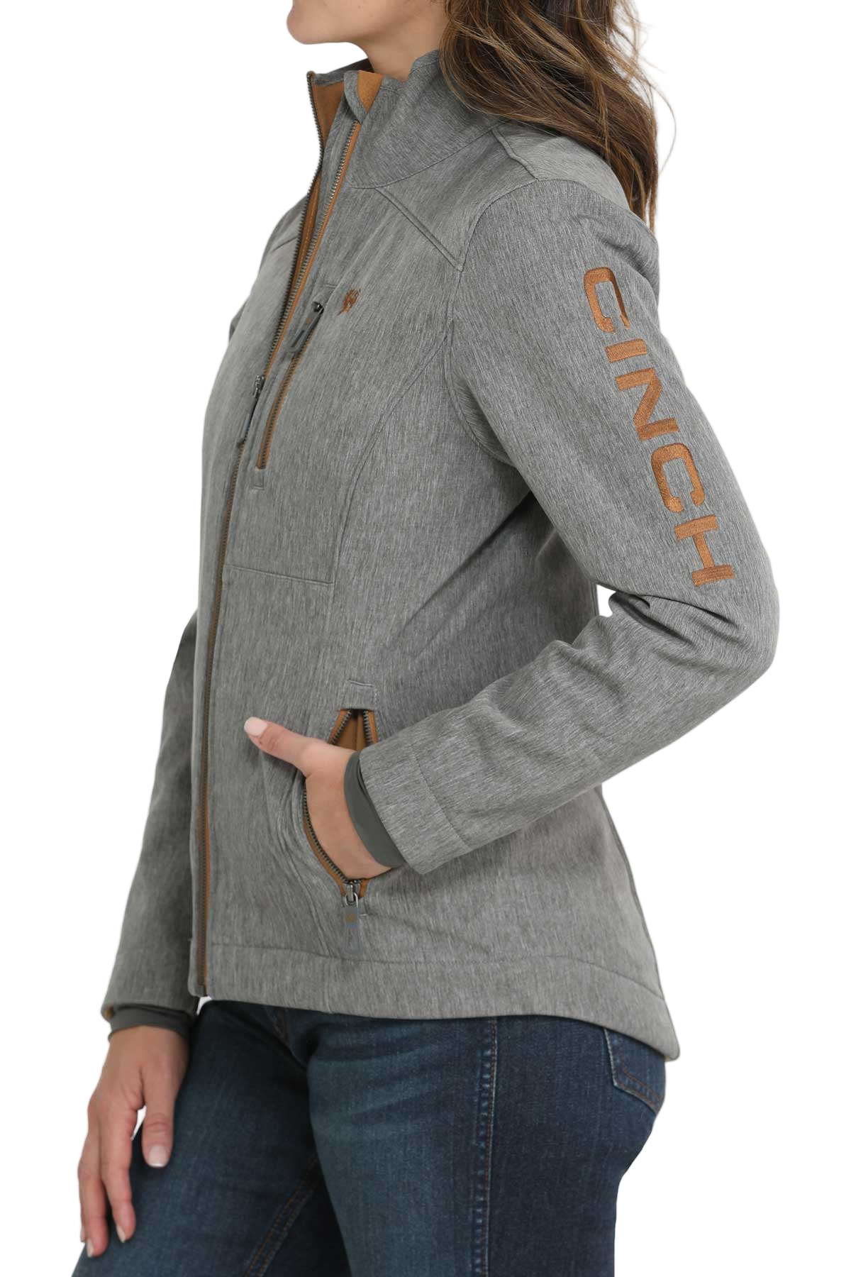 A woman wearing a light gray softshell jacket with brown zipper accents and “CINCH” printed down the sleeve, paired with dark blue jeans