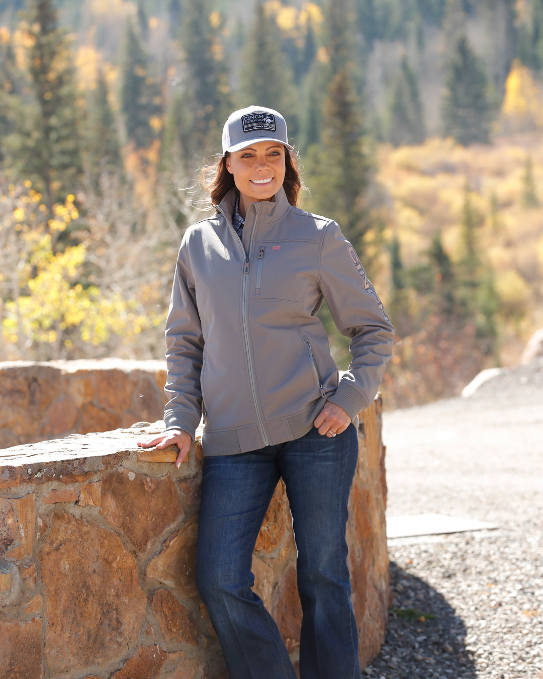 Woman wearing a gray jacket and cap standing outdoors with autumn trees in the background