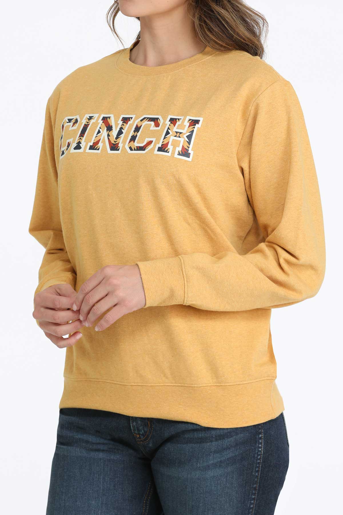 Woman wearing a gold Cinch crew neck sweatshirt with bold patterned CINCH logo and jeans on a white background