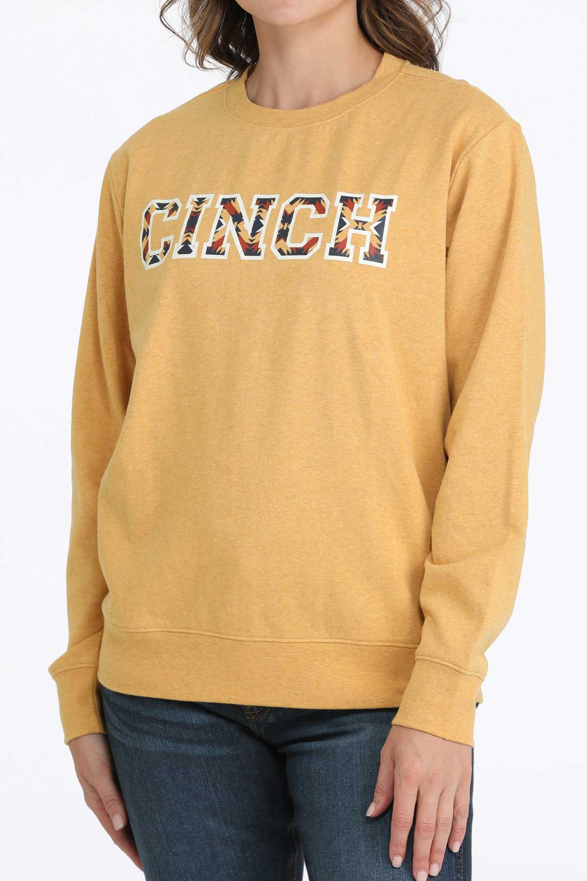 Woman wearing a gold Cinch crew neck sweatshirt with bold patterned CINCH logo and jeans on a white background