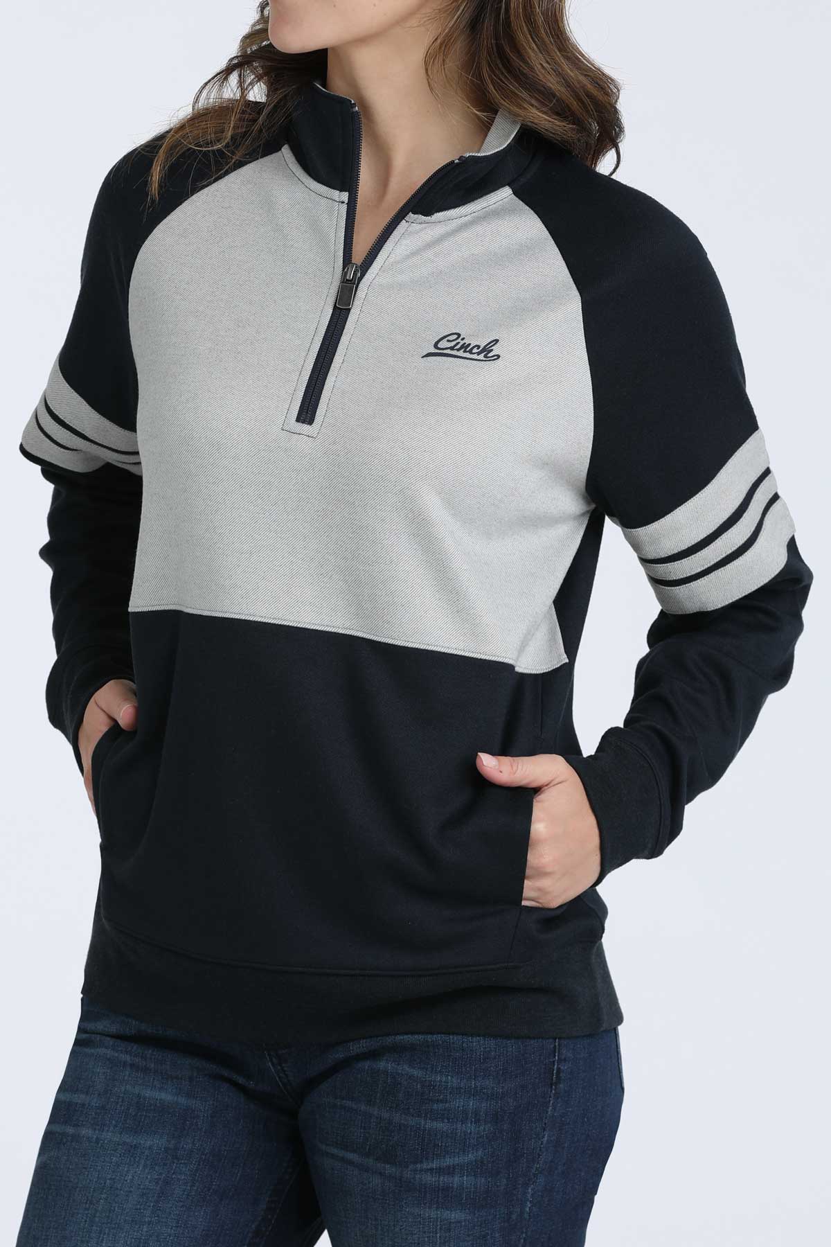 Woman wearing a navy and grey Cinch 1/4 zip sweatshirt with striped sleeves and jeans on a white background