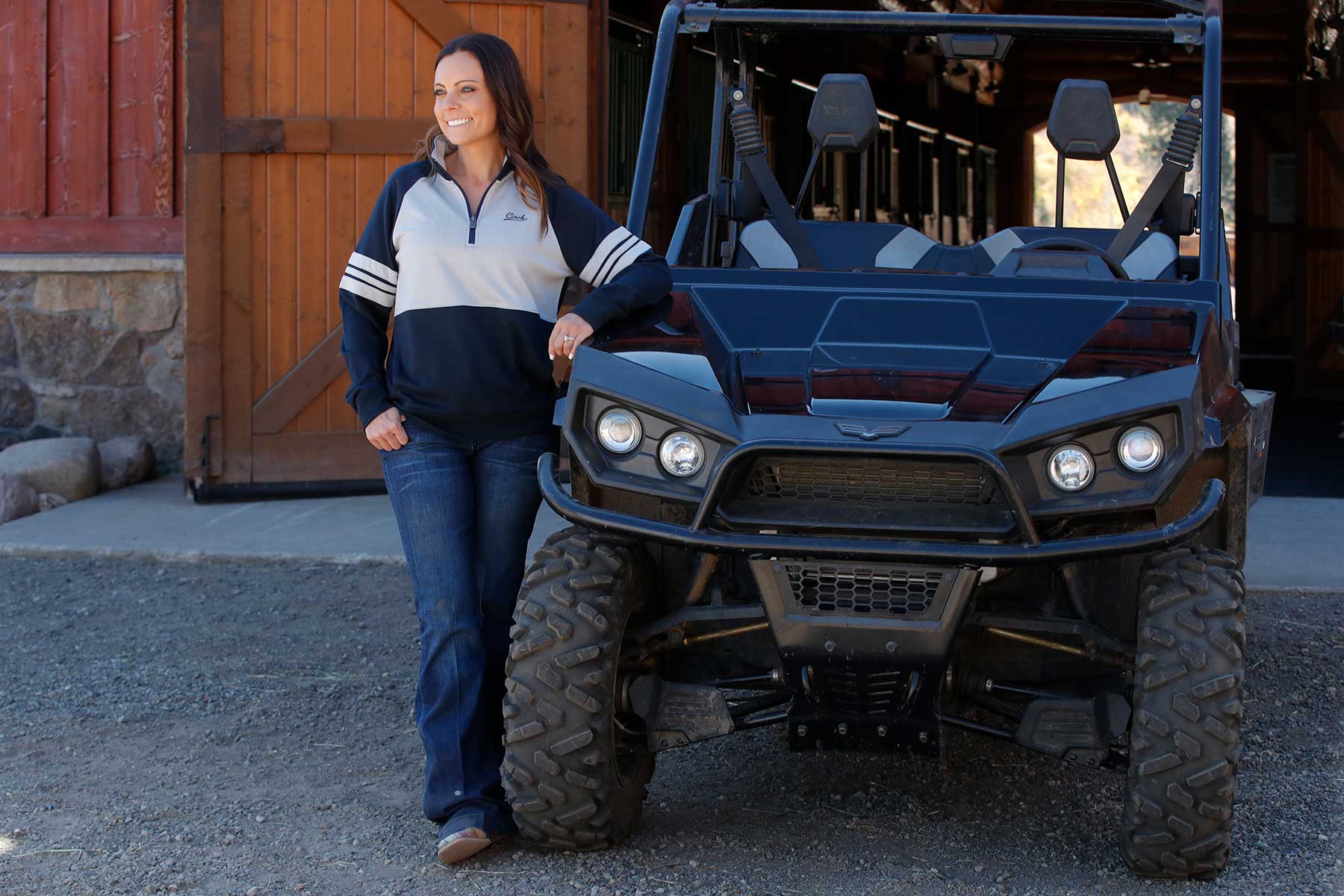 Outdoor image of woman wearing navy Cinch 1/4 zip sweatshirt with jeans, standing next to a utility vehicle