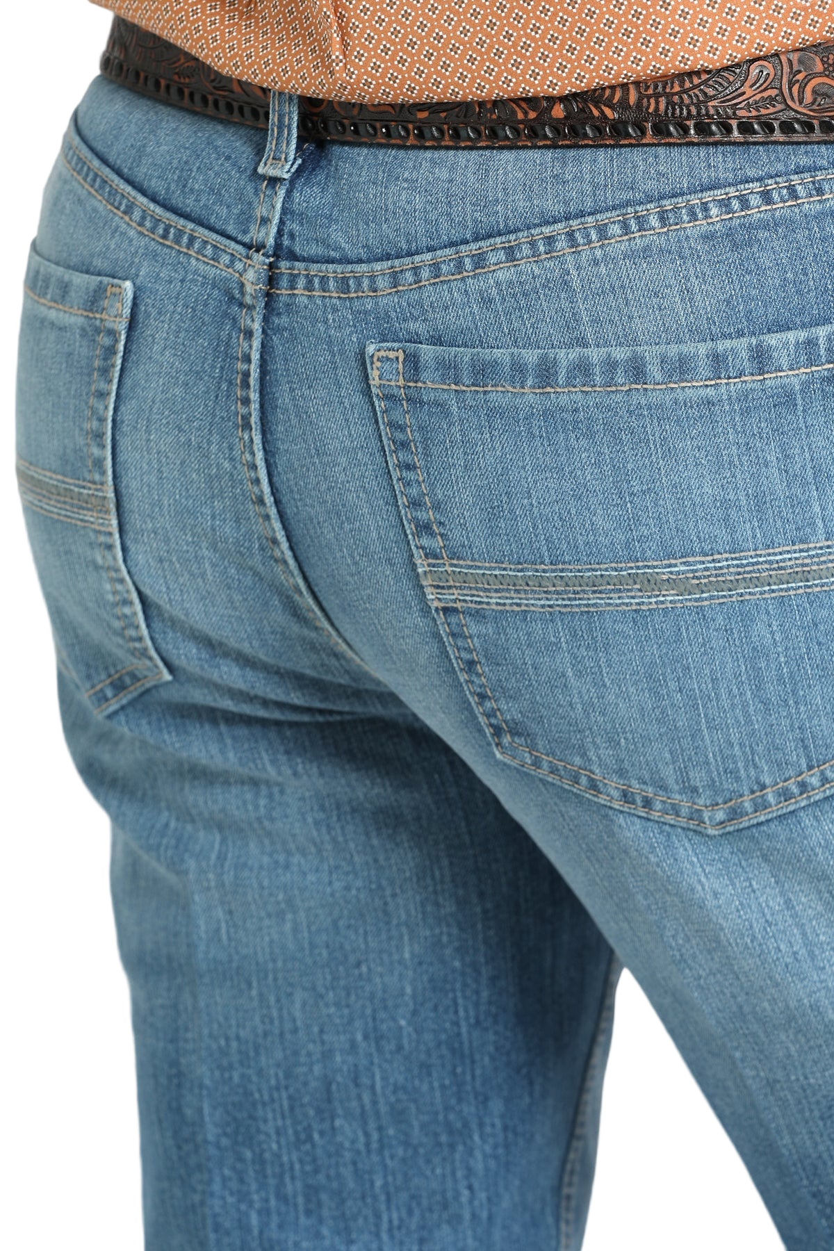 Close-up of the back of a person wearing blue denim jeans with simple stitching on the back pockets and a tooled brown leather belt, paired with a tucked-in patterned shirt