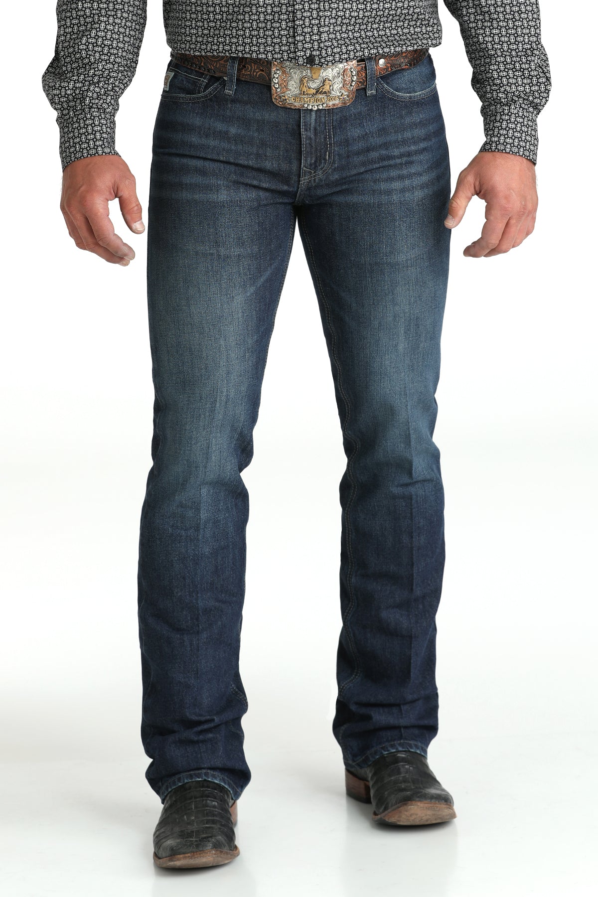 A man wearing dark wash jeans, a patterned shirt, a large belt buckle, and boots stands against a white background