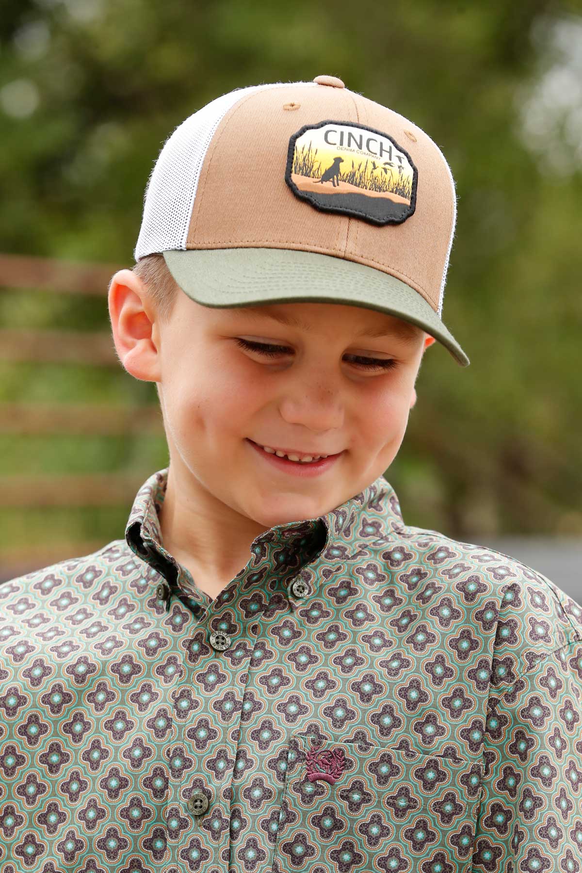 Close-up of a boy wearing a 'CINCH' hat and a collared button-down shirt standing outside