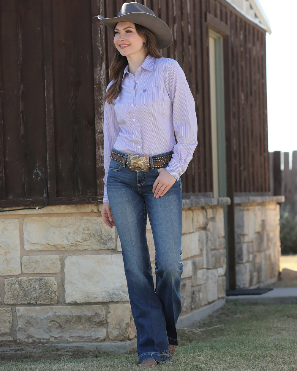 Woman wearing a cowboy hat, striped button-up shirt, jeans, and a large belt buckle, walking outside next to a rustic stone and wood building
