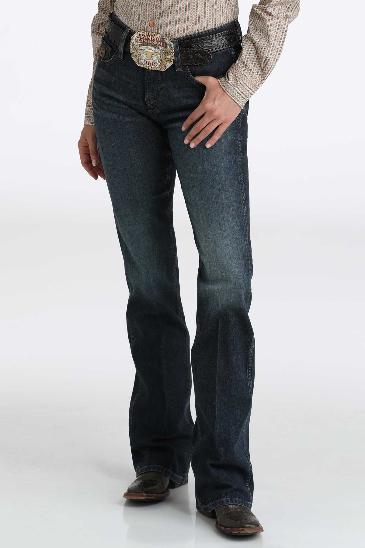 Woman wearing Cinch ladies’ dark wash boot cut jeans with a belt and boots on a white background