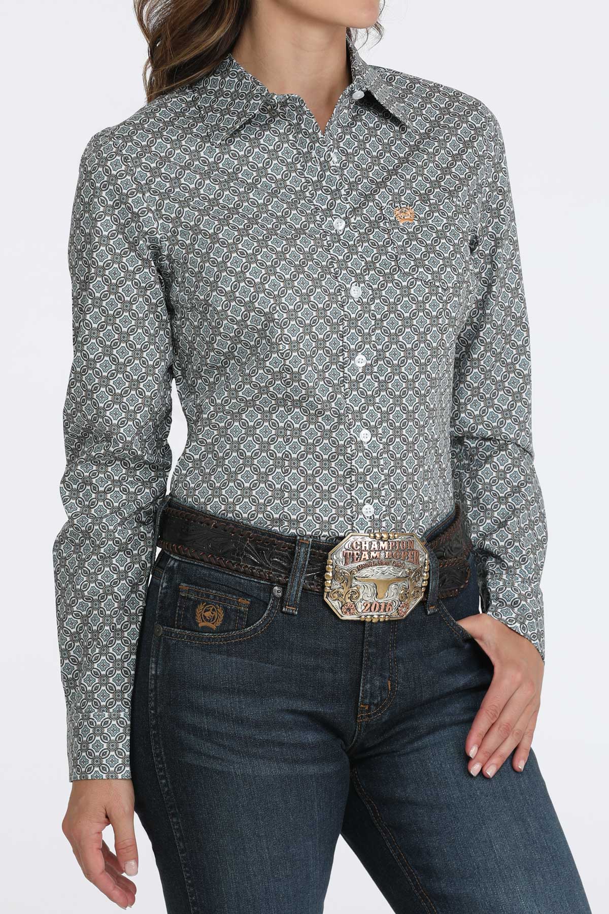 Woman wearing a Cinch ladies’ long sleeve white print shirt tucked into jeans with a large western belt buckle on a white background