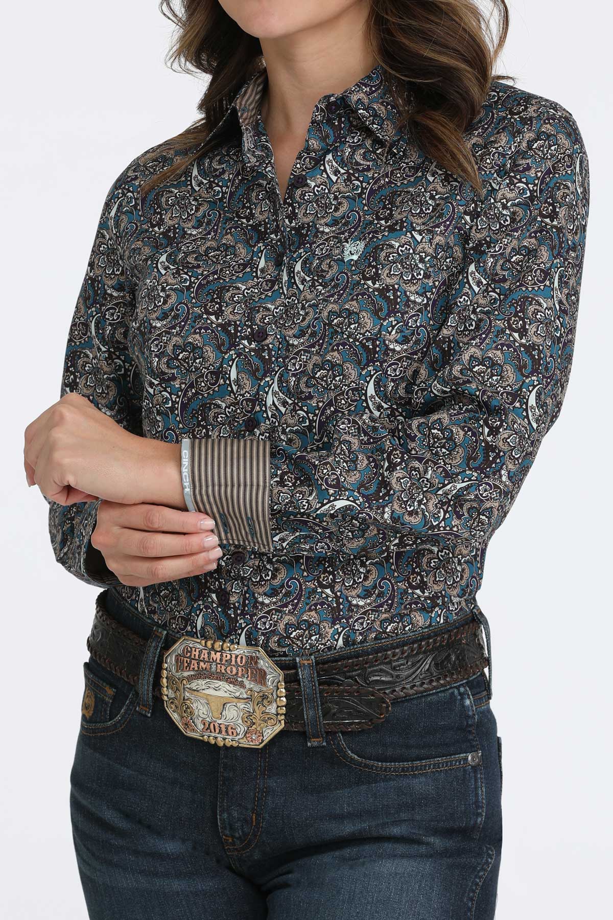 Woman wearing a dark paisley print button-up shirt with long sleeves, and a contrasting cuff detail, paired with dark jeans and a large belt buckle