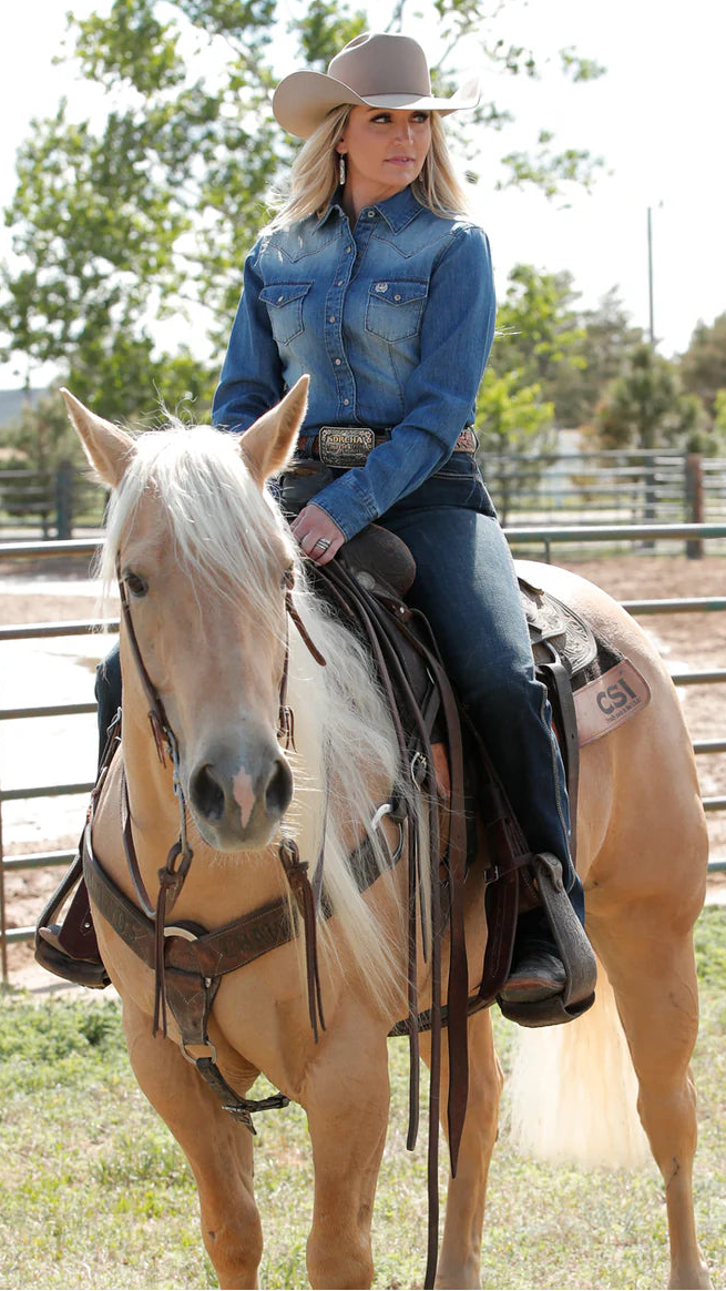 Woman wearing a denim shirt, jeans, and a cowboy hat, sitting on a light-colored horse in an outdoor arena