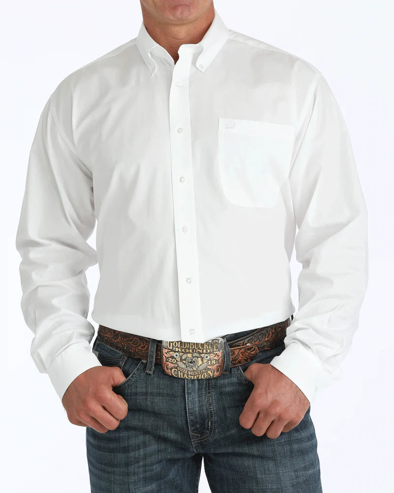 A man wearing a white button-up shirt tucked into dark jeans with a large belt buckle stands against a white background