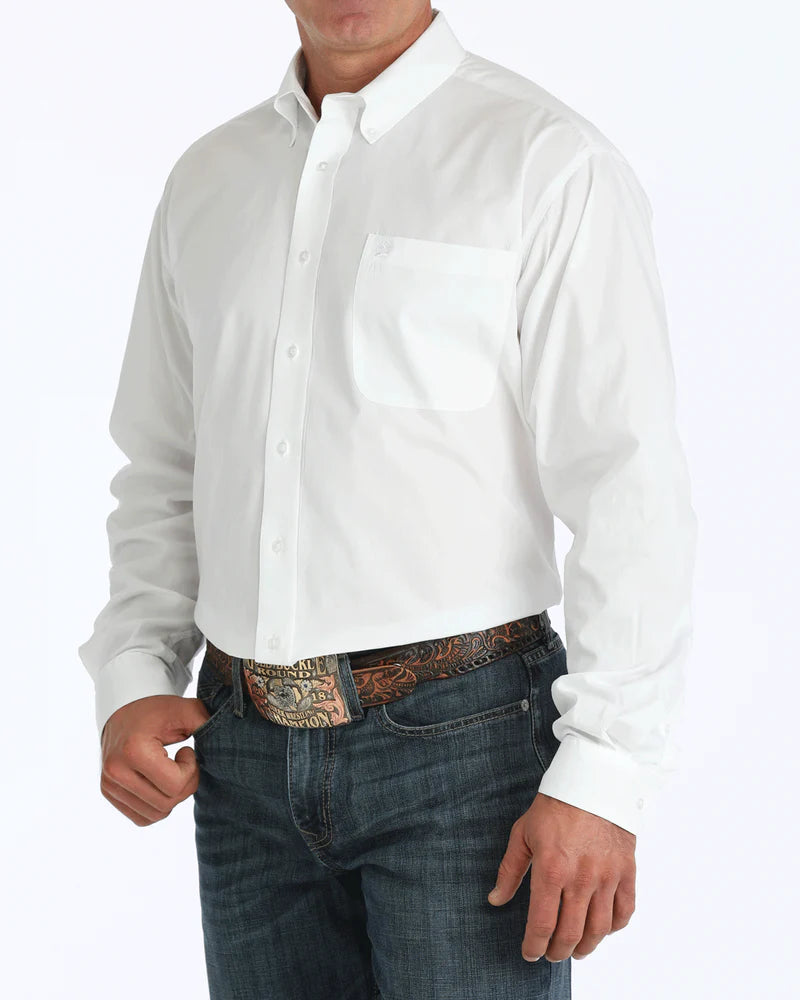 A man wearing a white button-up shirt tucked into dark jeans with a large belt buckle stands against a white background