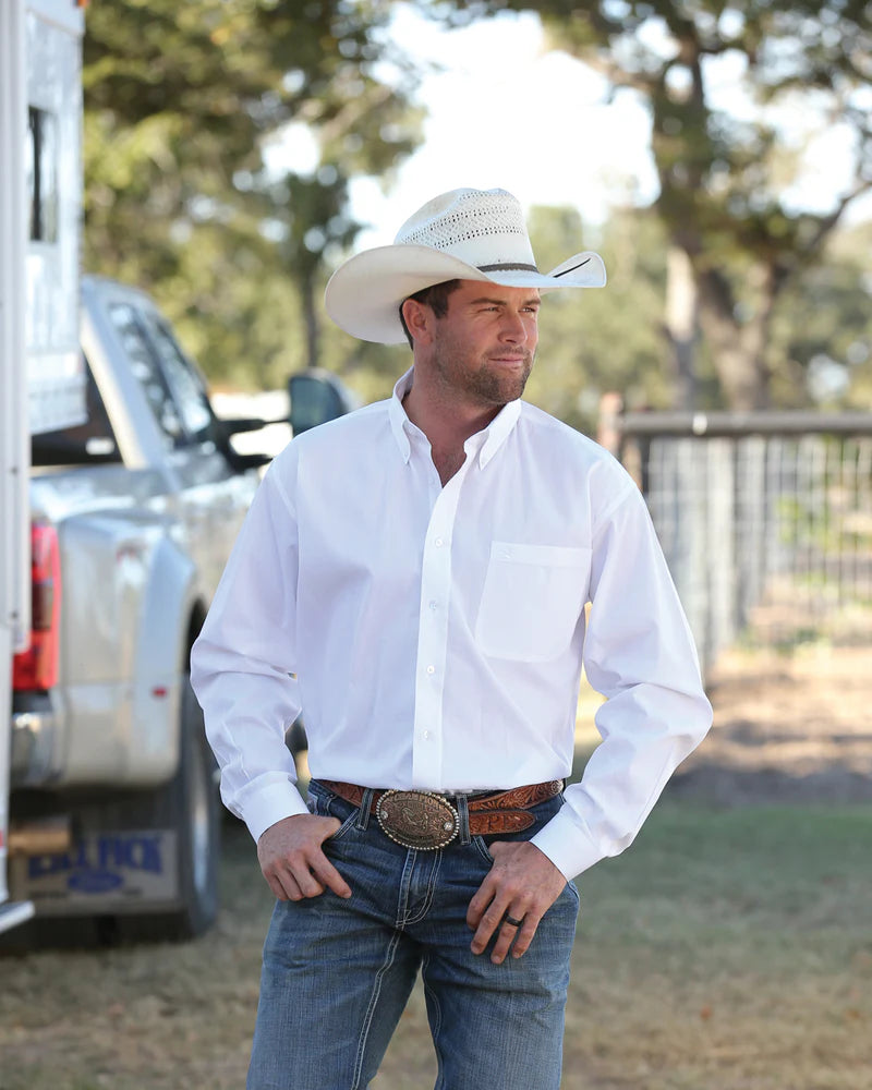 A man wearing a white button-up shirt, cowboy hat, jeans, and a large belt buckle stands outdoors near a truck