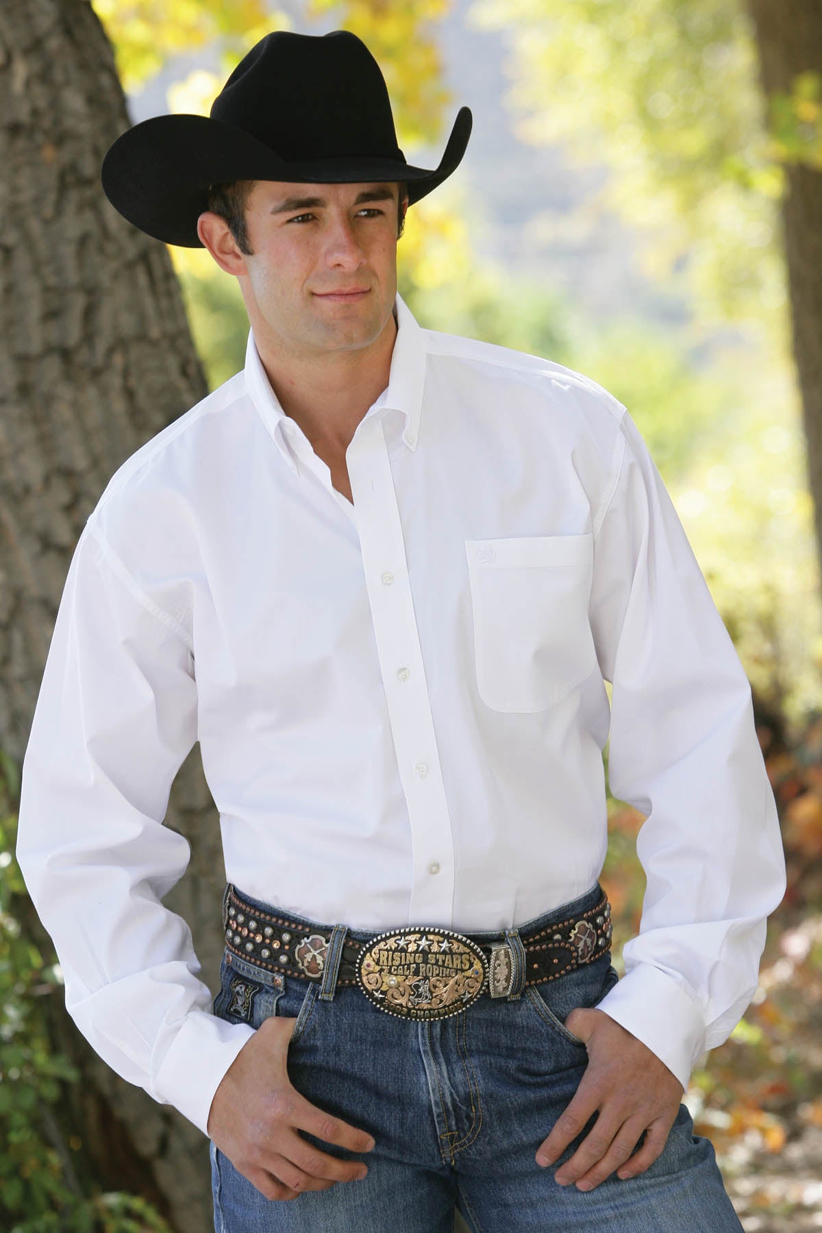 A man wearing a white button-up shirt tucked into dark jeans with a large belt buckle and a black cowboy hat stands outside in front of a tree