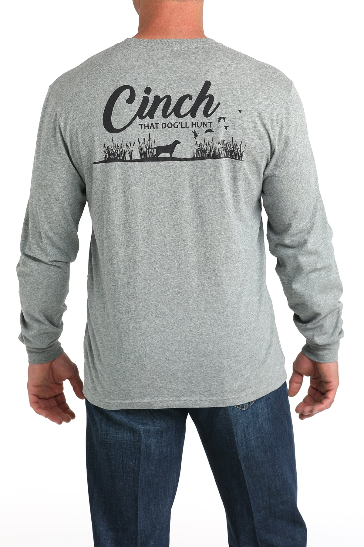 A man wearing a gray long-sleeve shirt and dark jeans is shown from the back. The shirt has a black graphic with the word “Cinch” and a hunting dog design