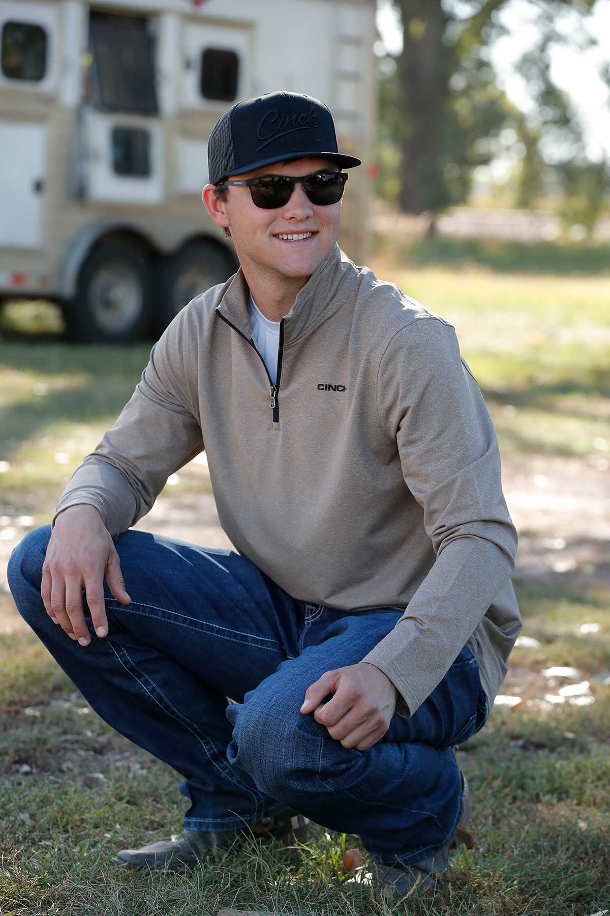 Man in a beige jacket and blue jeans squatting outdoors with a blurred background