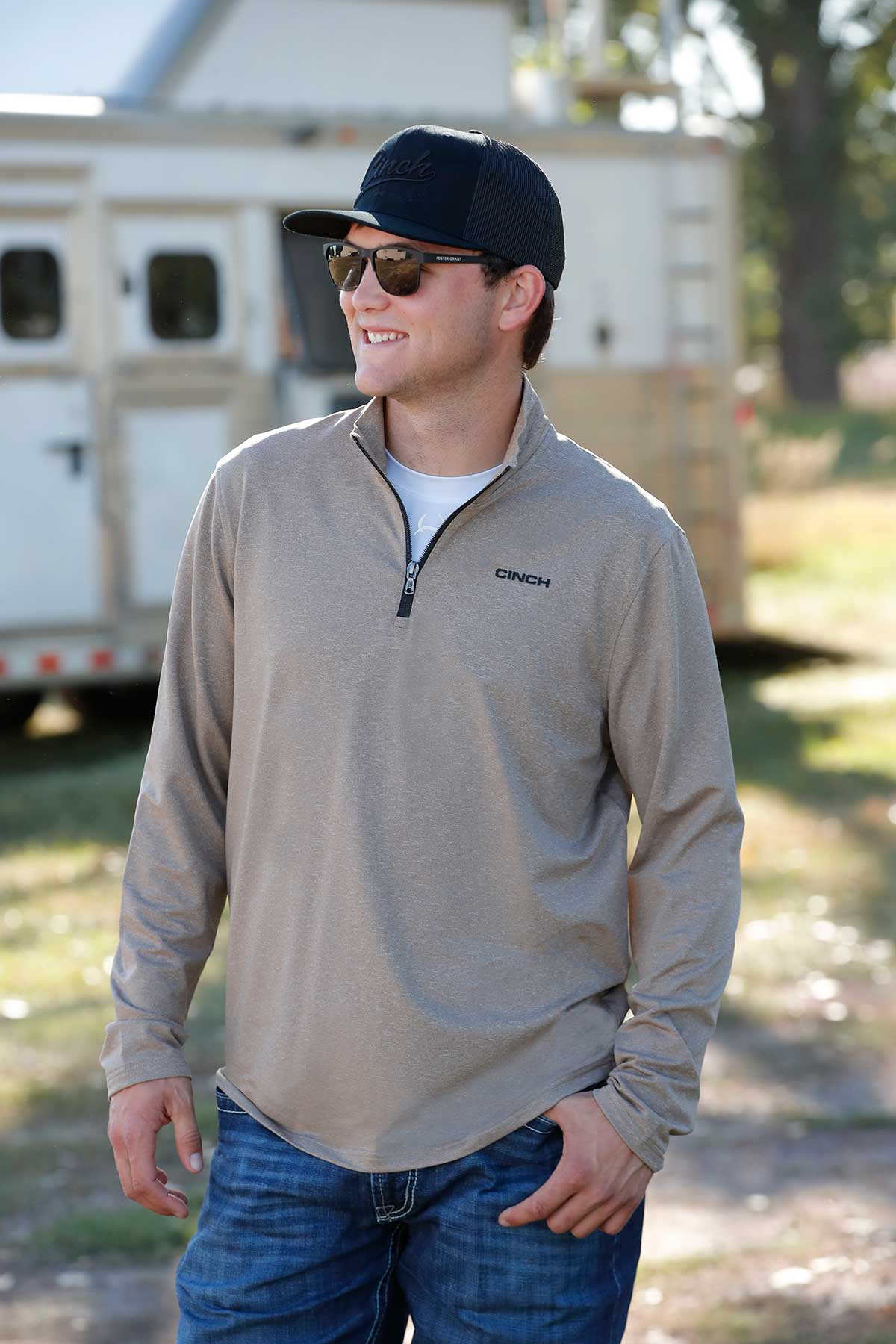 Man in a beige jacket and blue jeans standing outdoors with a blurred background