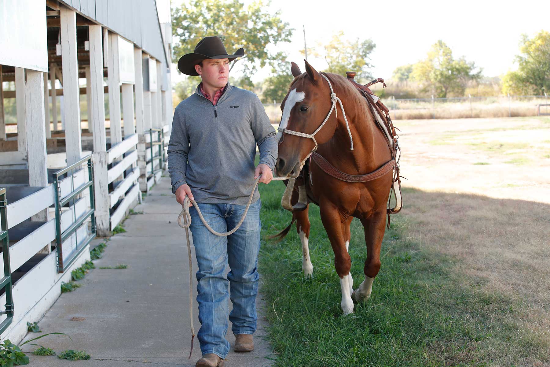Man wearing a gray quarter-zip pullover, blue jeans, and a cowboy hat standing next to a brown and white horse outside a barn
