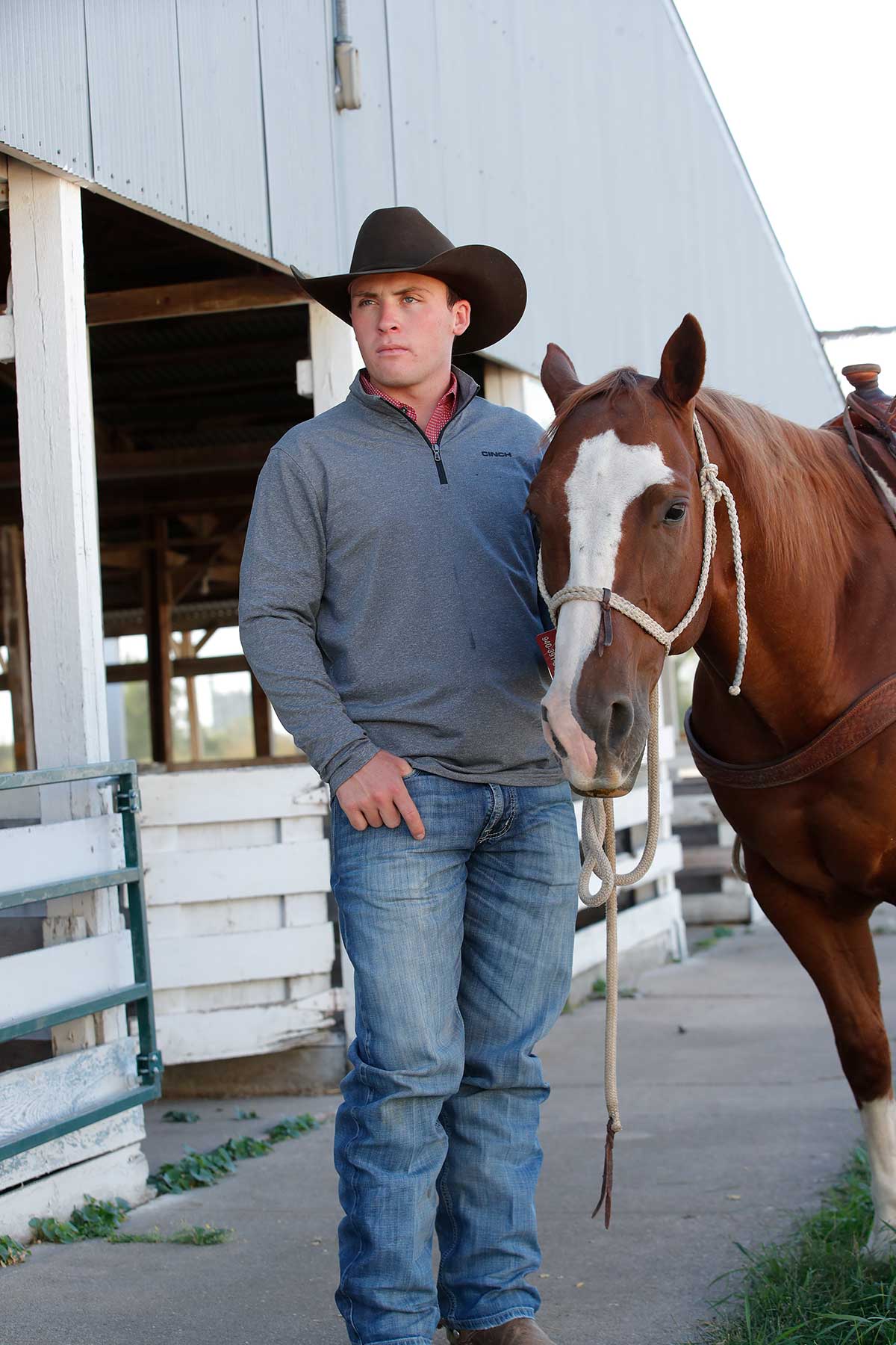 Man wearing a gray quarter-zip pullover, blue jeans, and a cowboy hat standing next to a brown and white horse outside a barn