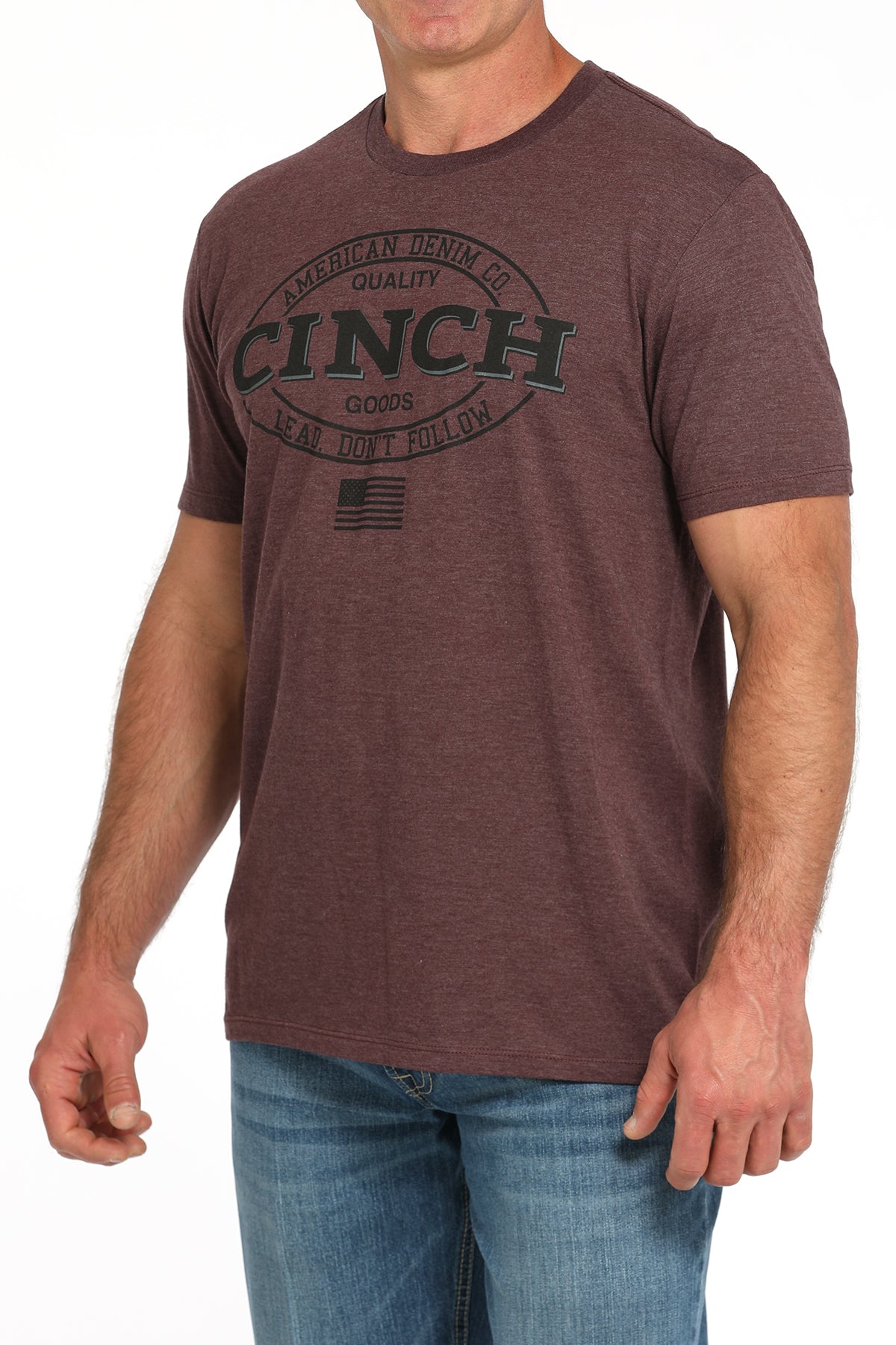Man wearing a brown t-shirt with a large 'Cinch' graphic on the front and blue jeans on a white background
