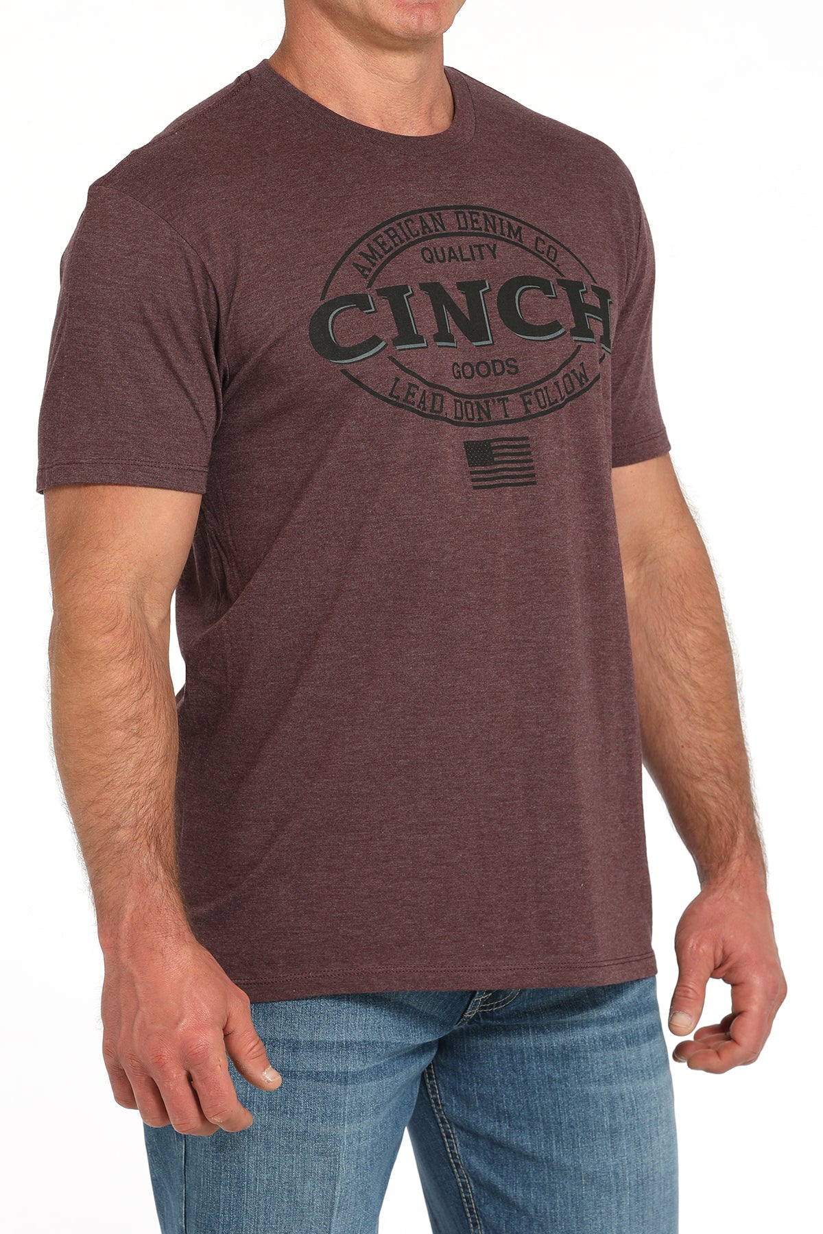 Man wearing a brown t-shirt with a large 'Cinch' graphic on the front and blue jeans on a white background