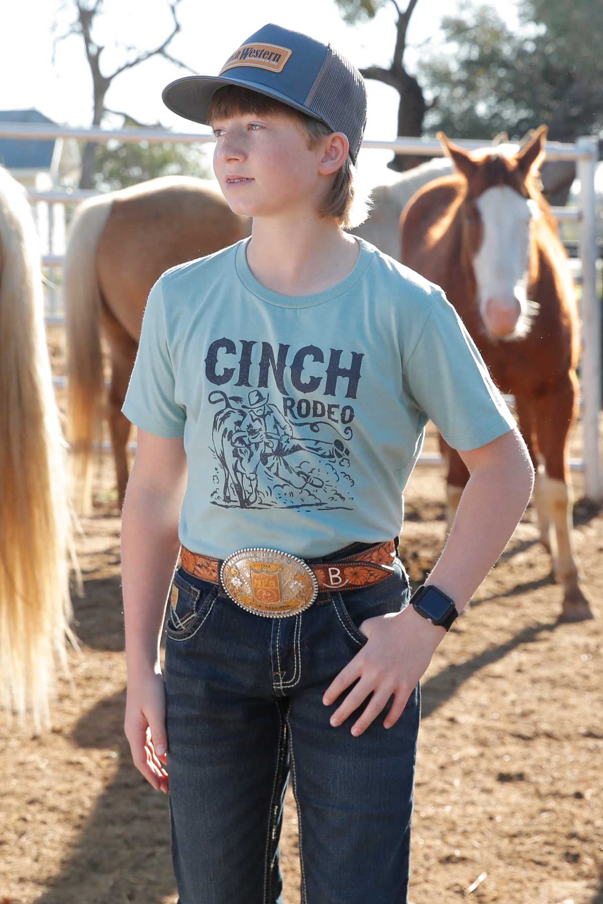 Boy standing outside in front of horses wearing a green 'CINCH' t-shirt with jeans, a belt, and a hat