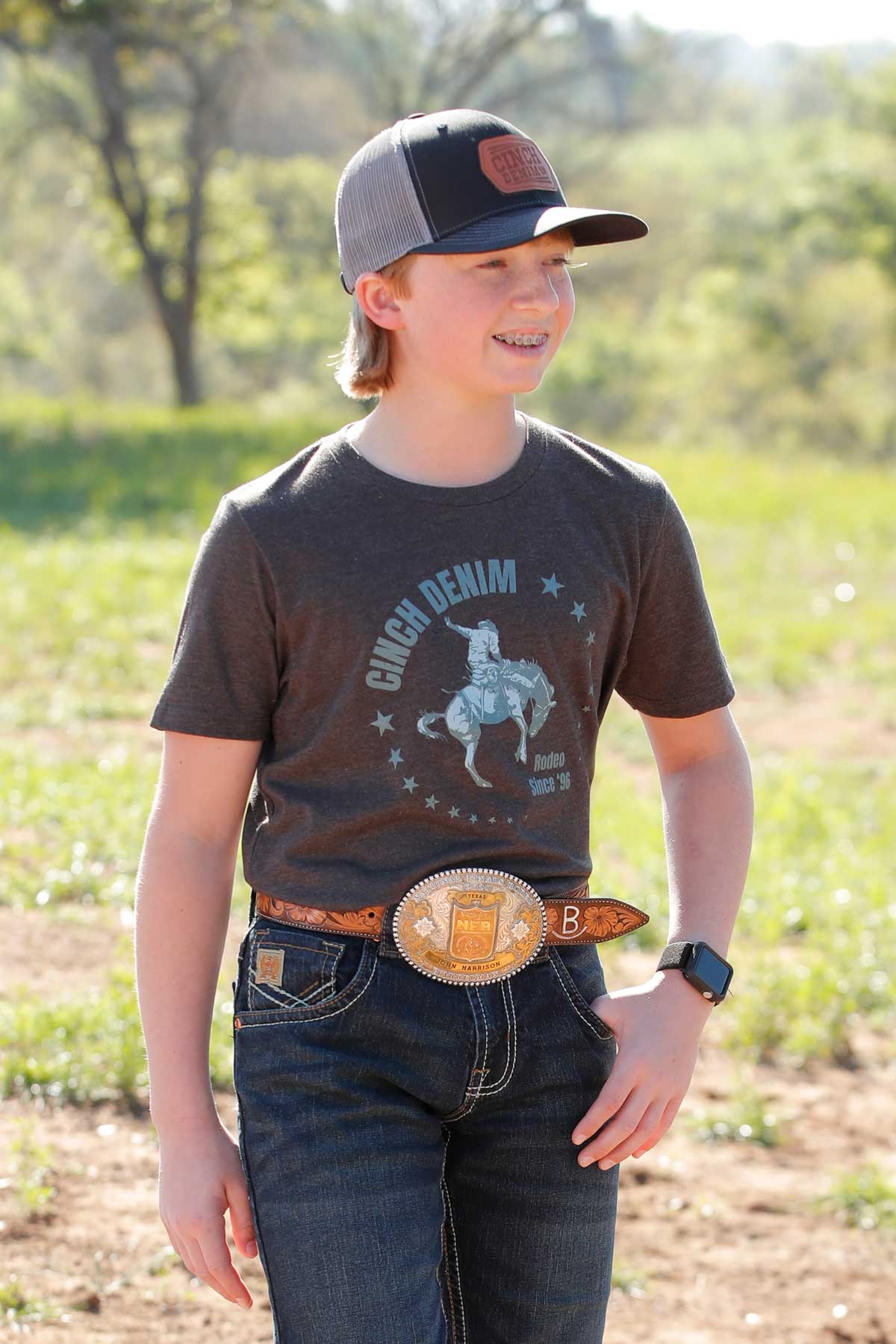 Boy standing outside wearing a gray 'CINCH' t-shirt with jeans, a belt, and a hat