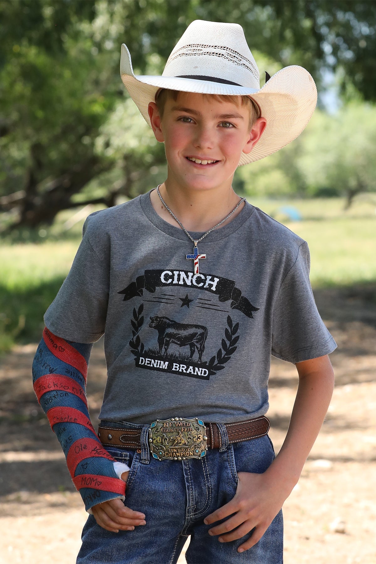 Boy standing outside wearing a gray t-shirt with jeans, a belt, and a cowboy hat with his right arm in a cast