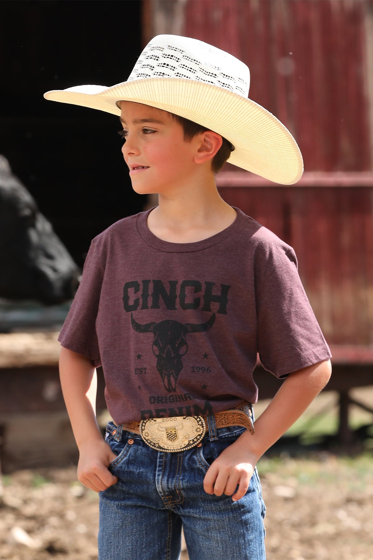 Boy standing outside wearing a maroon t-shirt with jeans, a belt, and a cowboy hat