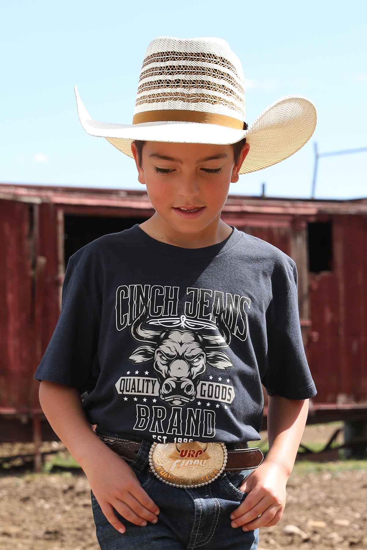 Little boy standing outside wearing a gray t-shirt with jeans, a belt, and a cowboy hat